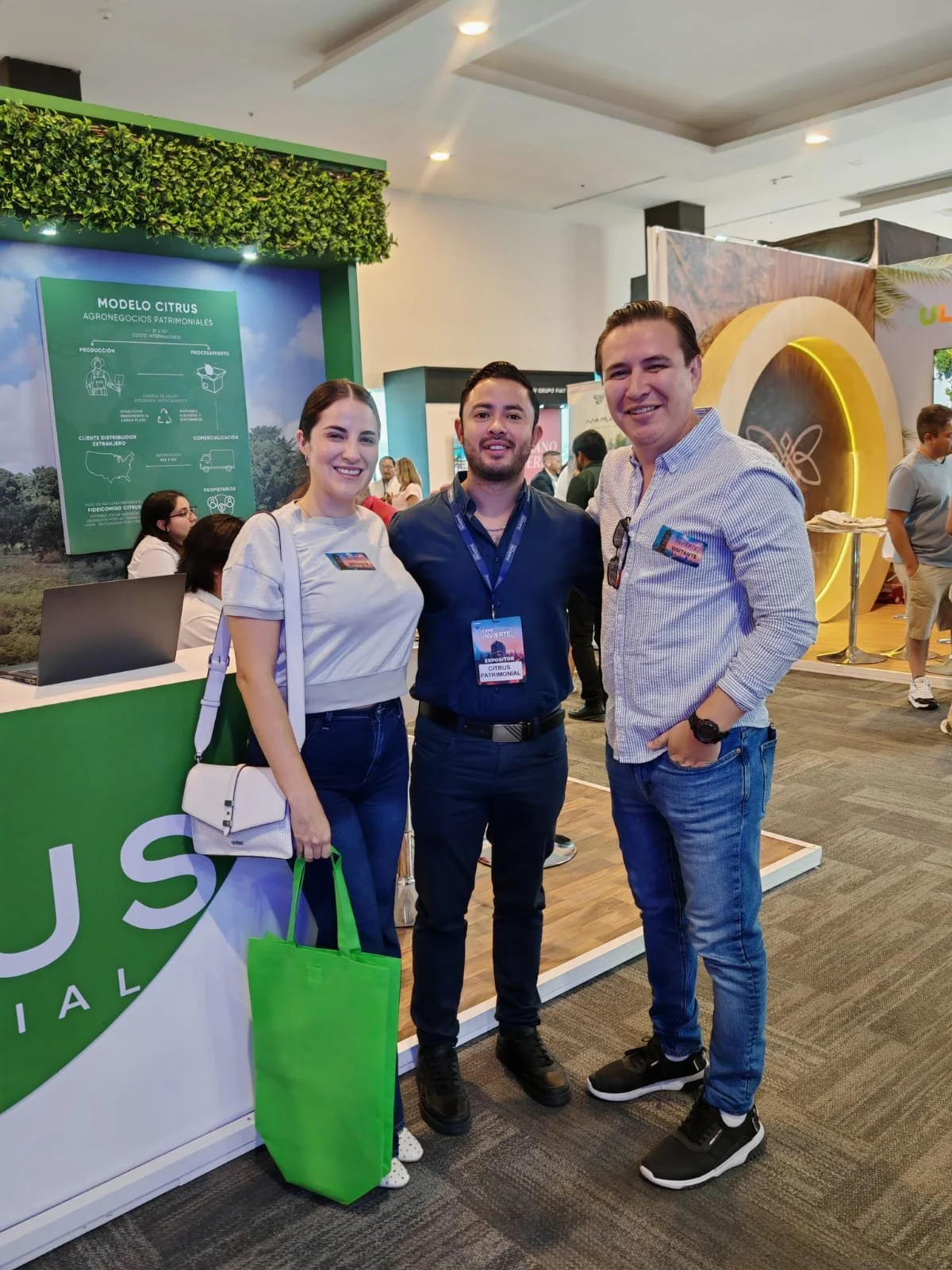 Tres personas en un evento, posando y sonriendo frente a un stand con señalización y decoración verde, en un espacio interior con muchas personas.