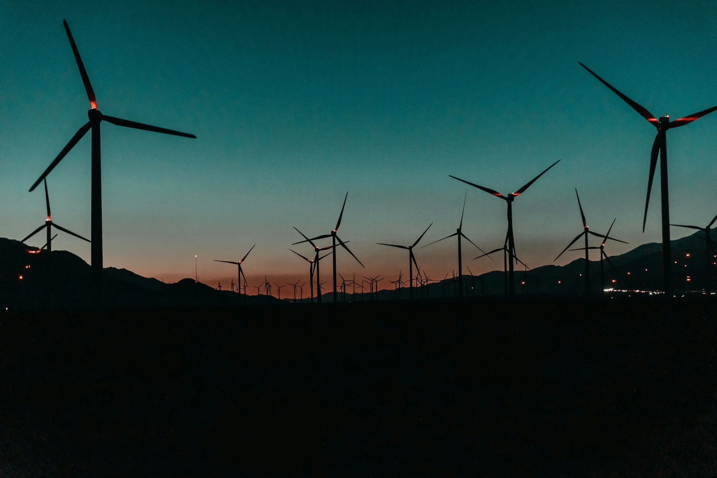 A row of wind turbines during dusk with a mountain range in the background and a colorful sky.