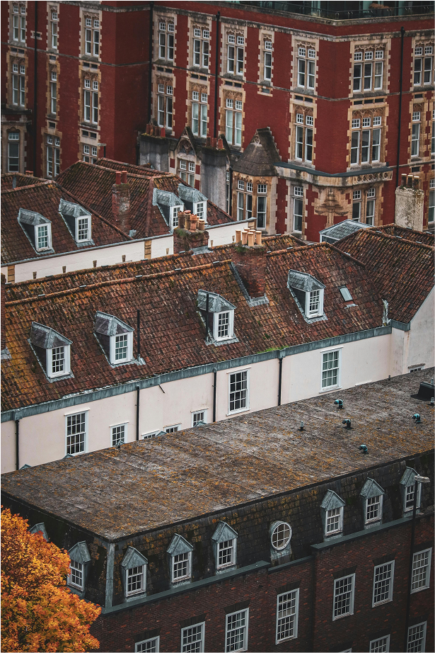 A view of rooftops of old buildings with red brick and white walls, some with dormer windows and chimneys, in a cityscape.