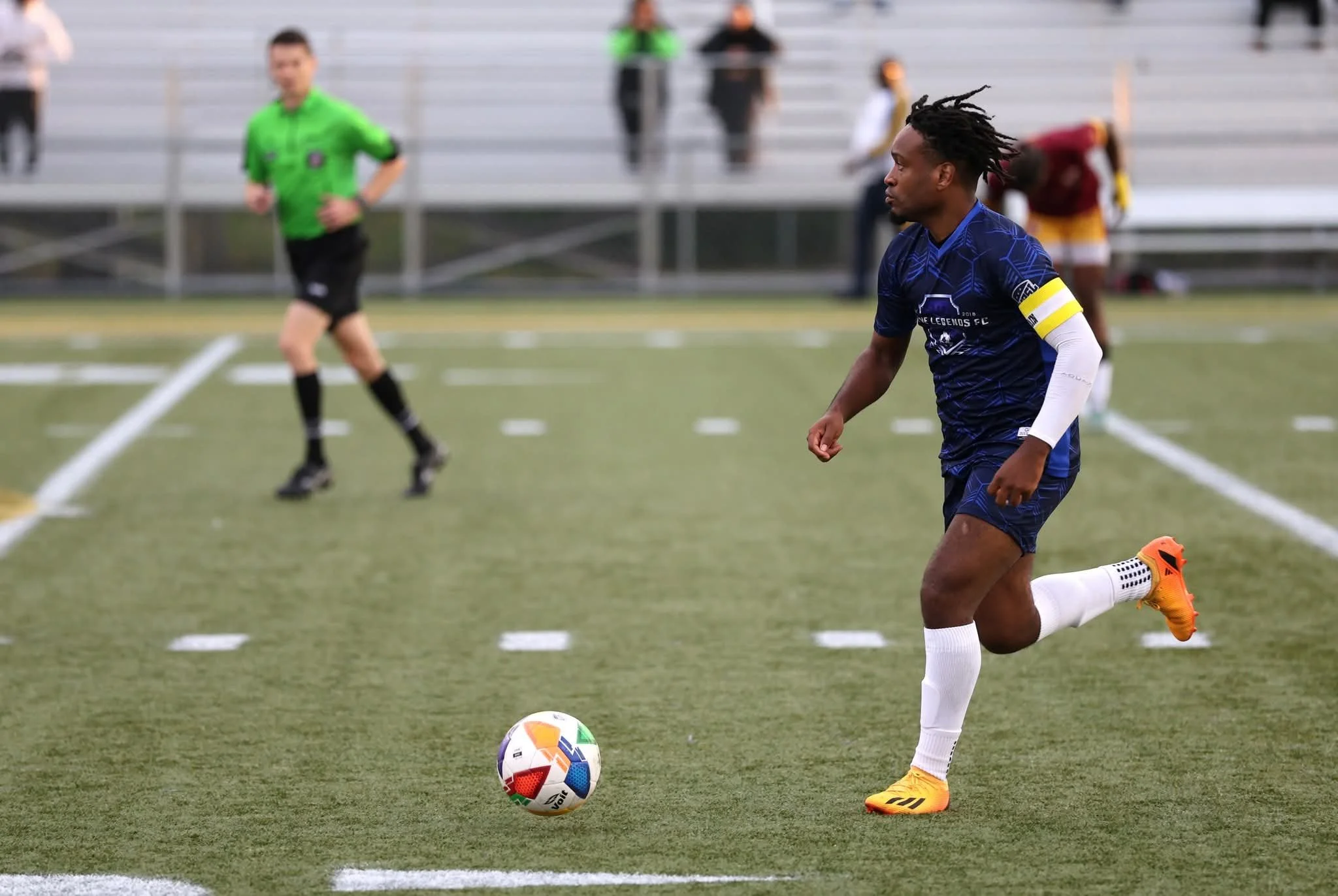 A soccer player in blue uniform and orange shoes running on the field with a soccer ball, with a referee and other players in the background.