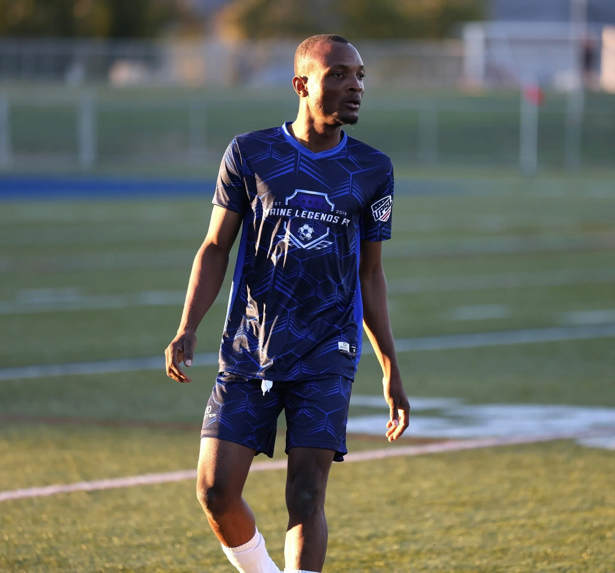 A man wearing a blue soccer uniform with the logo 'Main Legends FC' stands on a soccer field during sunset.