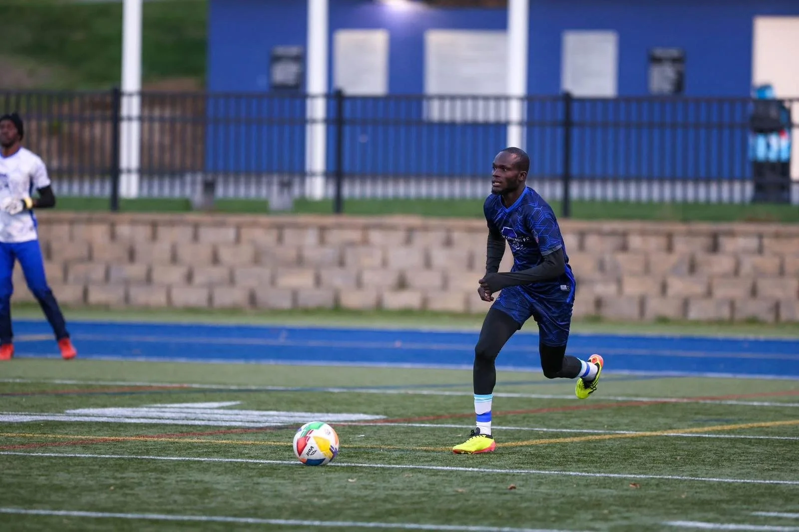 A soccer player in a blue uniform is preparing to kick a soccer ball on a field, with another player in the background and a blue building behind a black fence.