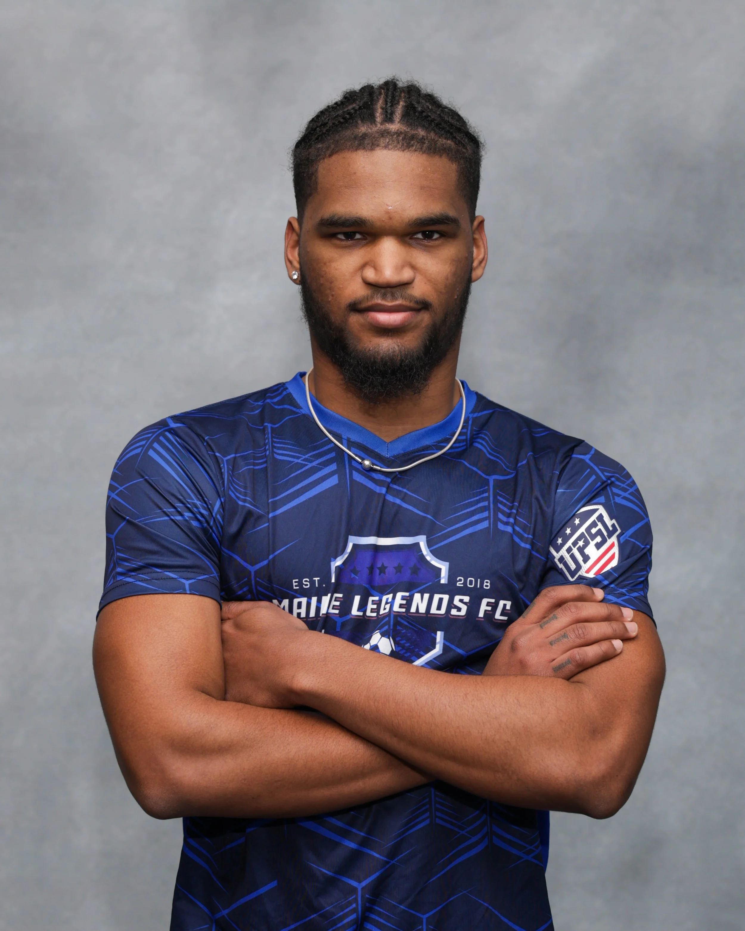 A soccer player standing on a field in a blue team uniform with white socks and purple shoes, with empty bleachers in the background.