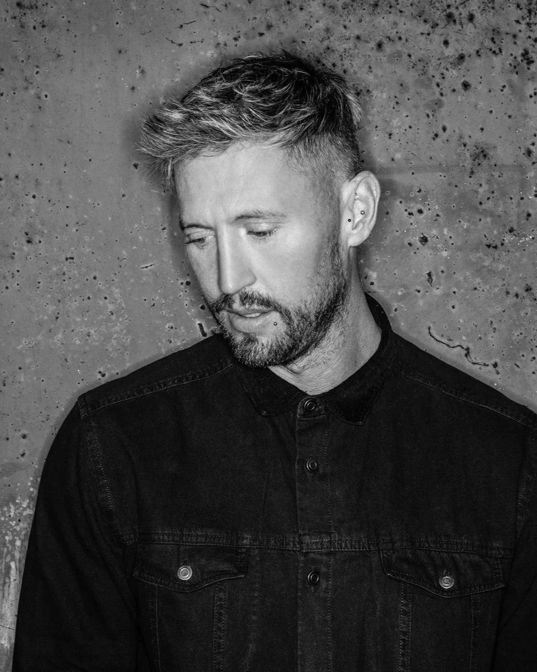 Cristoph. Black and white portrait of a man with short, styled hair and a beard, looking down, wearing a dark denim jacket, against a speckled concrete wall background.