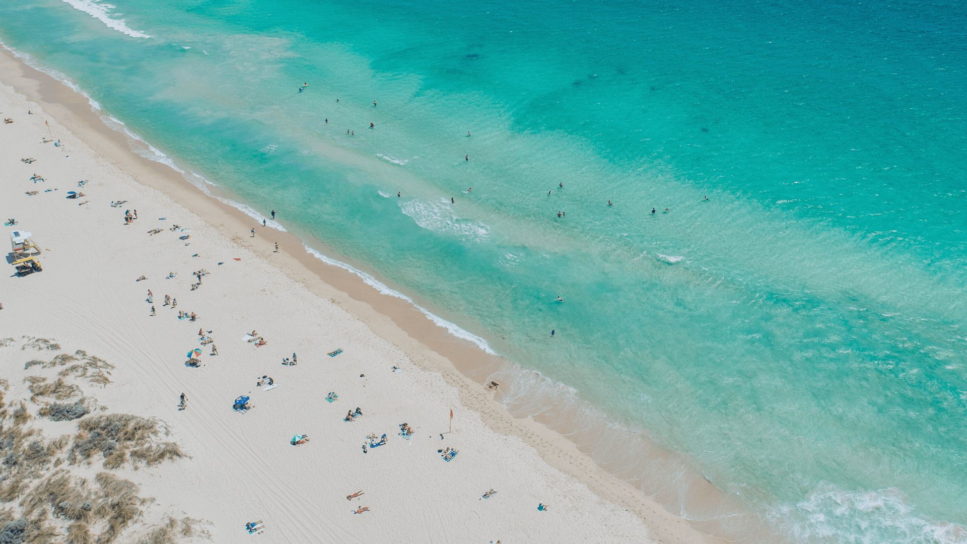 Electric Island Adelaide 2026 Event. Aerial view of a beach with white sand, turquoise water, and people sunbathing and swimming.