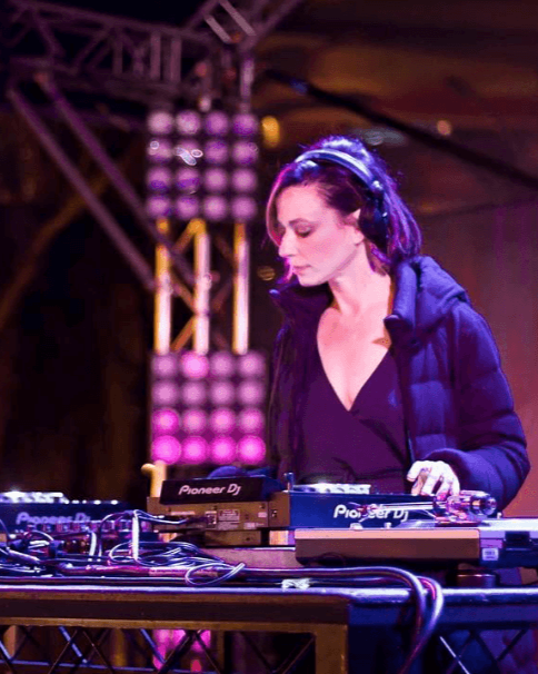 Felicity. A female DJ wearing headphones and a dark jacket, operating DJ equipment at a live event.
