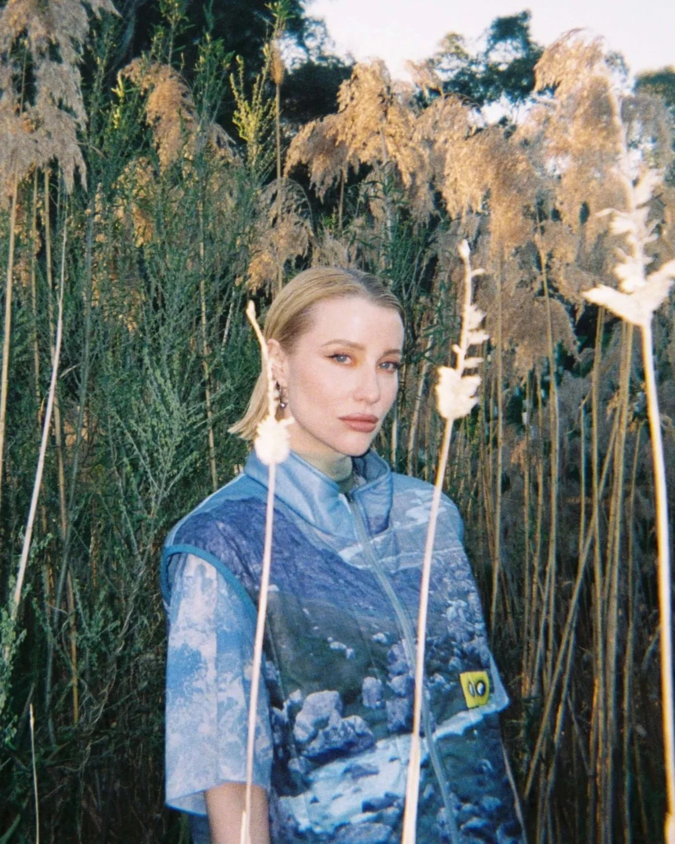 Rromarin. A young woman with blonde hair and earrings standing in a field of tall, beige dried plants or grasses during sunset.