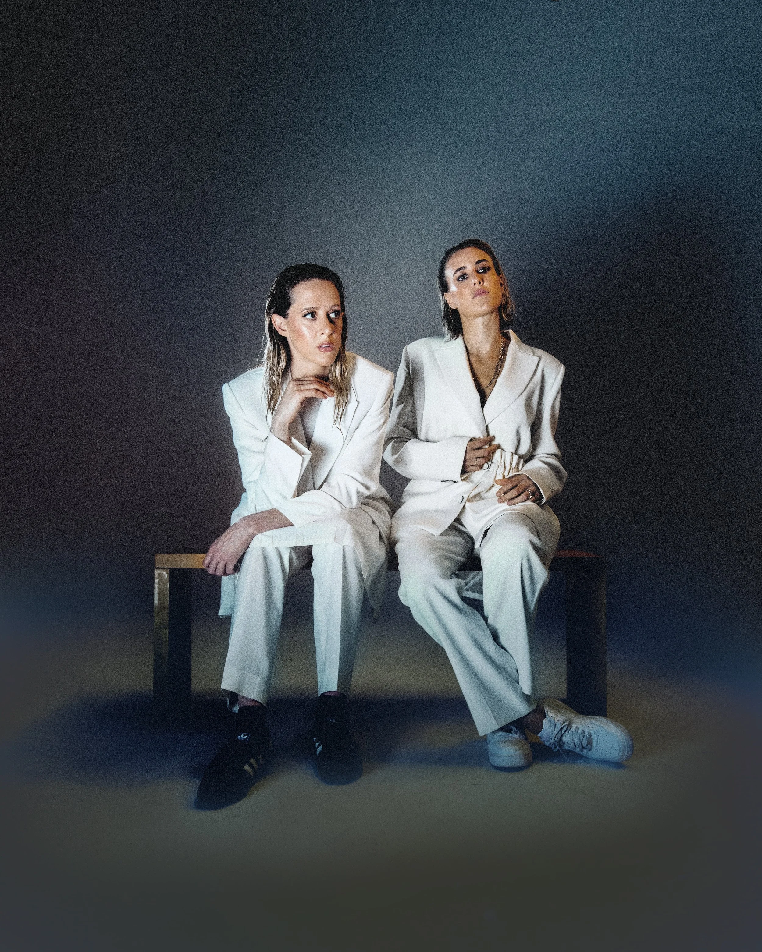 Eli & Fur. Two women dressed in white suits sitting on a bench against a dark background.