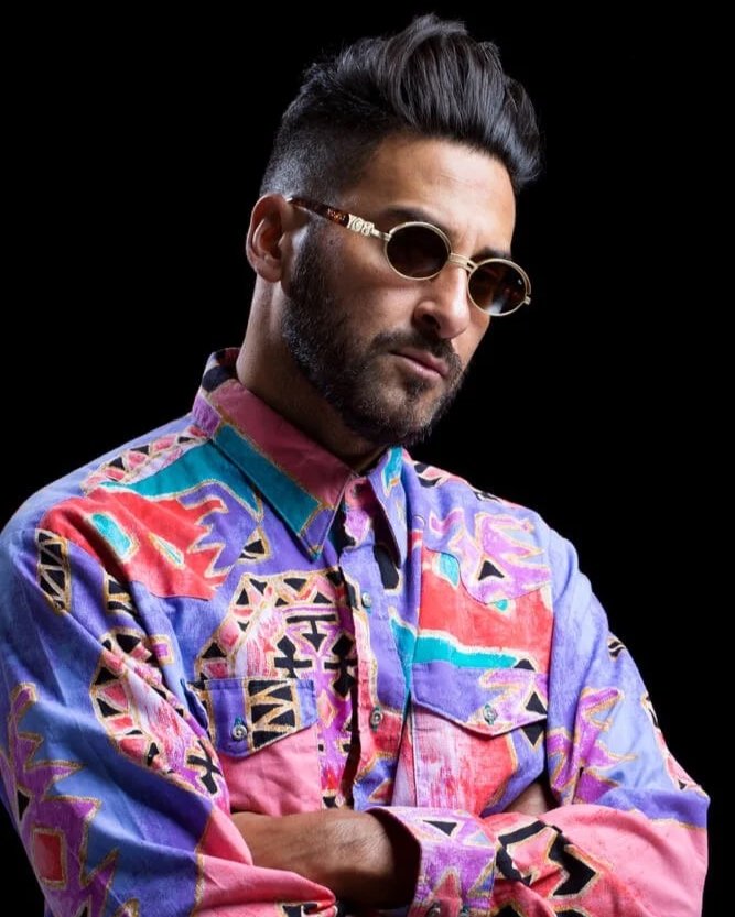 Armand van Helden. A man with dark hair styled upward, wearing dark sunglasses and a colorful, patterned shirt with pink, purple, blue, and red hues, standing against a black background.