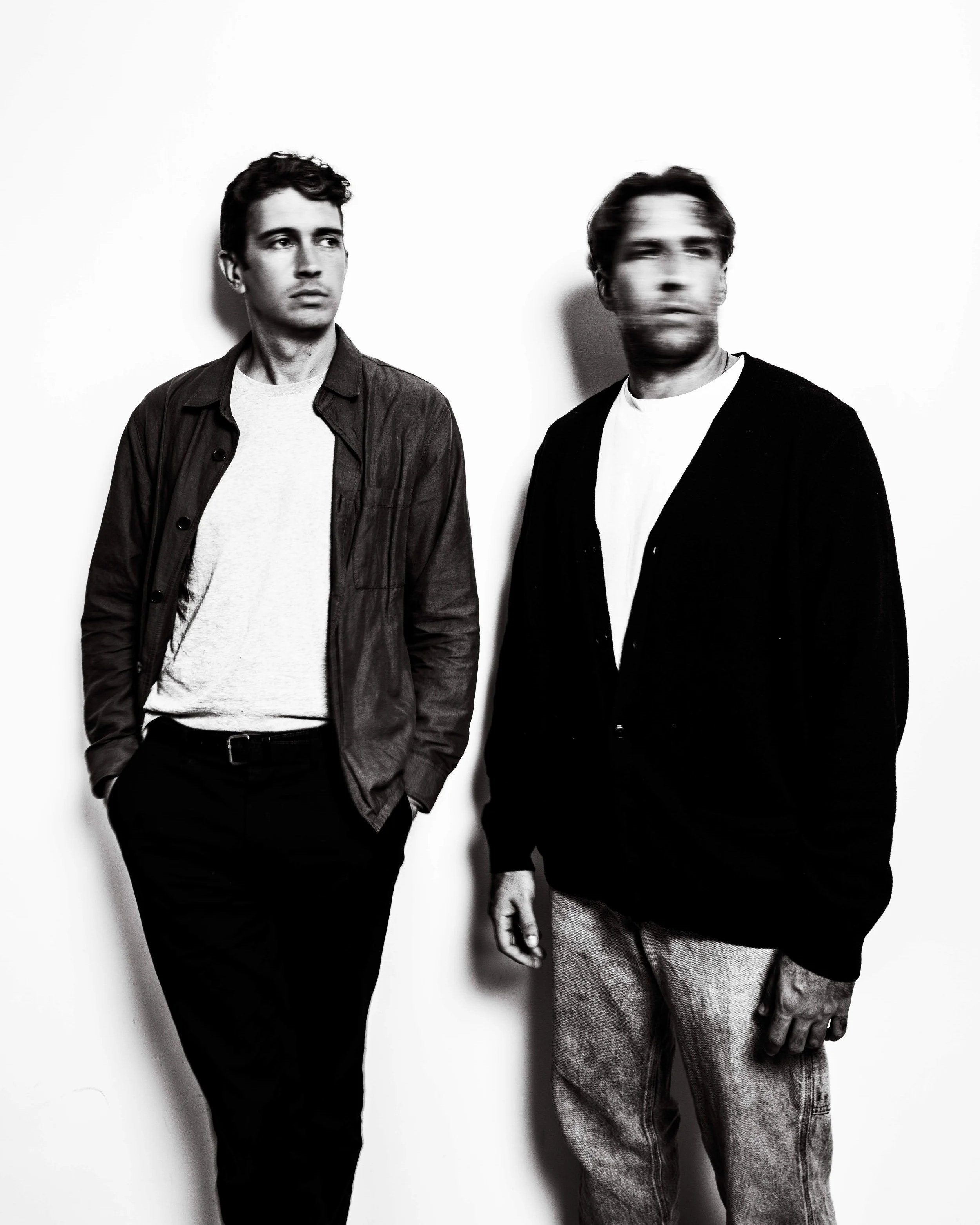 Flight Facilities (DJ Set). Two men standing against a plain white wall, one on the left looking to the right and the other on the right with a blurred face, dressed casually in dark and light clothing.