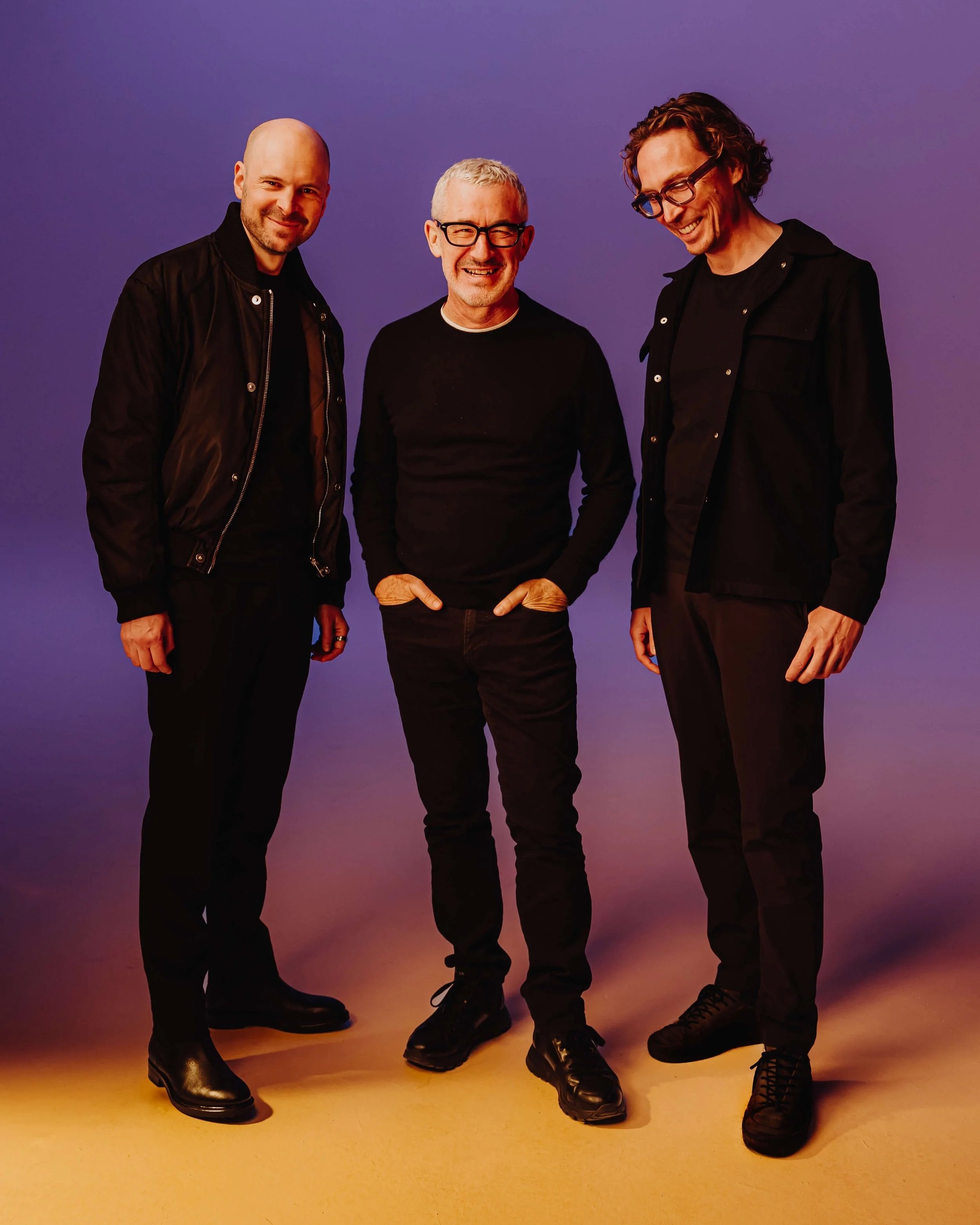 Above and Beyond. Three men standing together against a colorful background, smiling and dressed in black clothing.