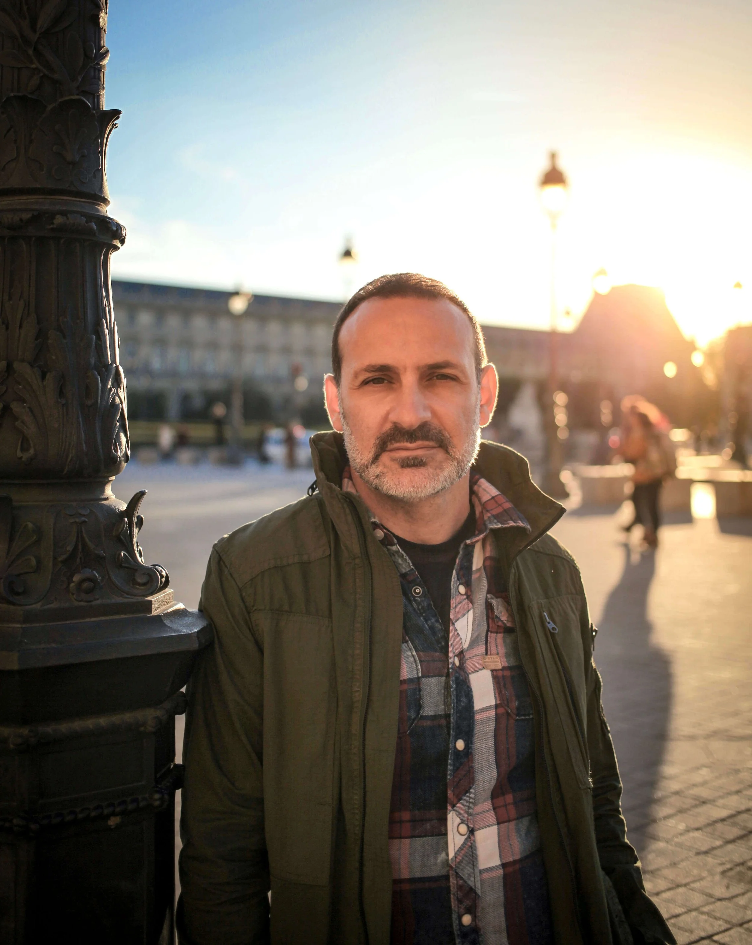 Late Night Tuff Guy. A man standing outdoors at sunset, leaning against a decorative black iron lamppost. He has short dark hair, a beard, and is wearing a green jacket over a plaid shirt.