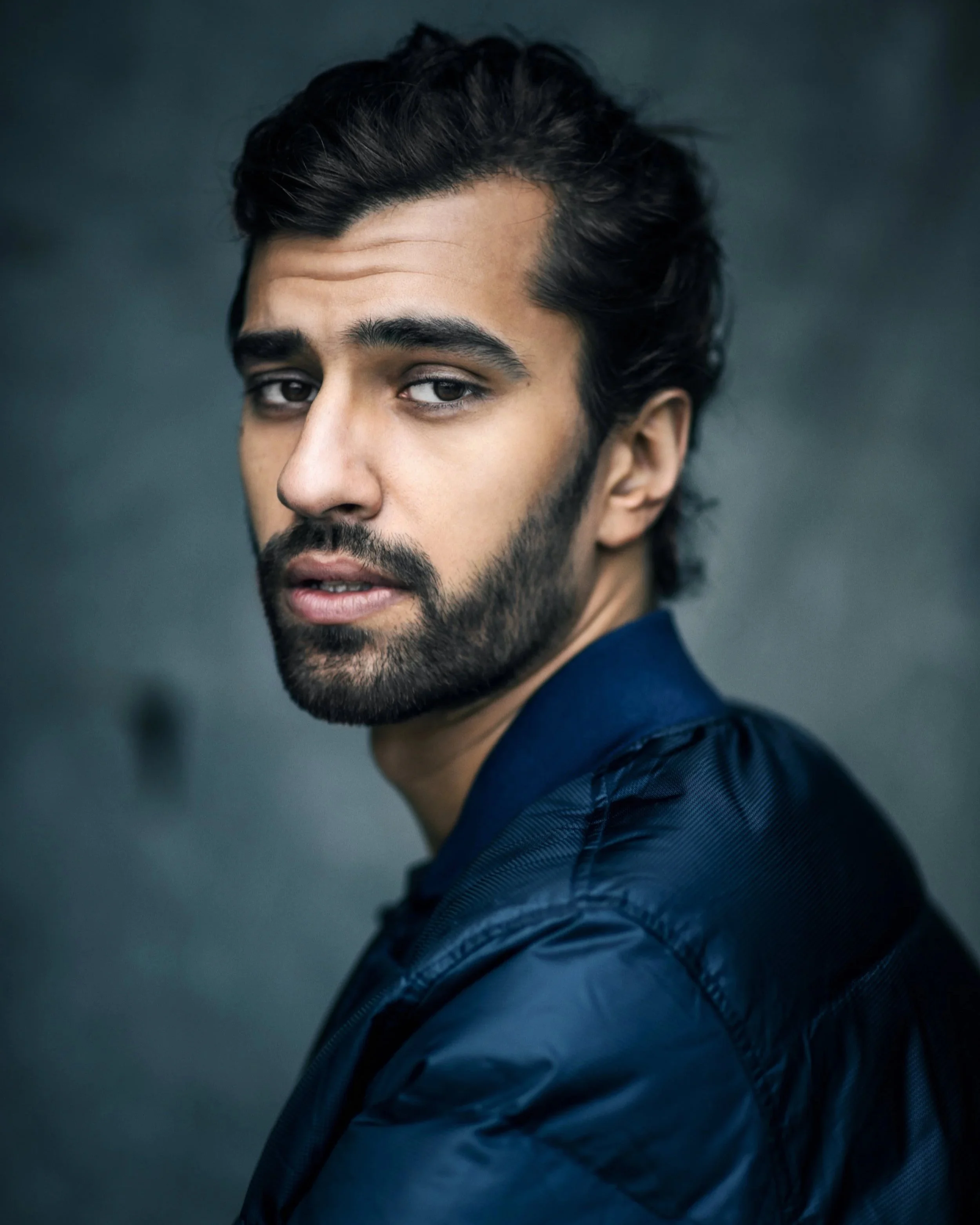Jeremy Olander. A young man with dark hair and a beard looking over his shoulder at the camera against a dark background.
