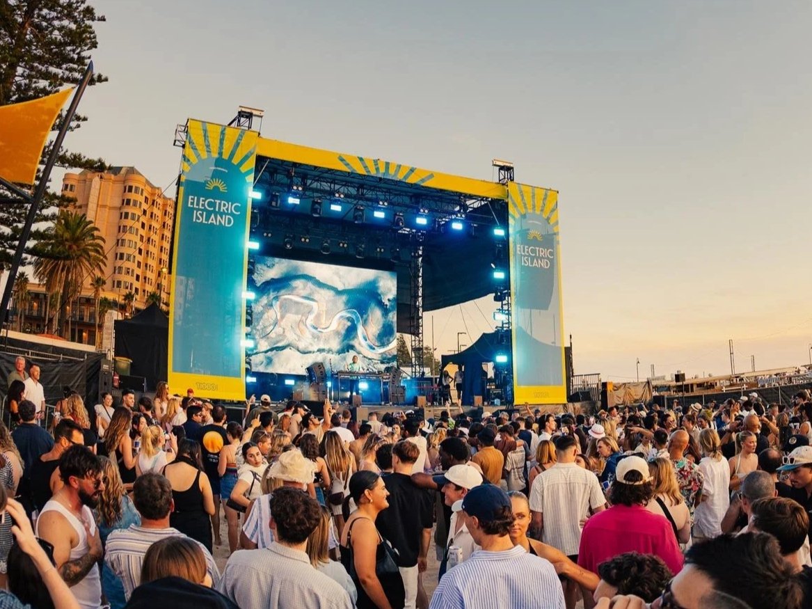 Electric Island Adelaide 2026 Event. Crowd of people attending a concert on an outdoor stage with a large screen and banners labeled "Electric Island" during sunset in an urban setting.
