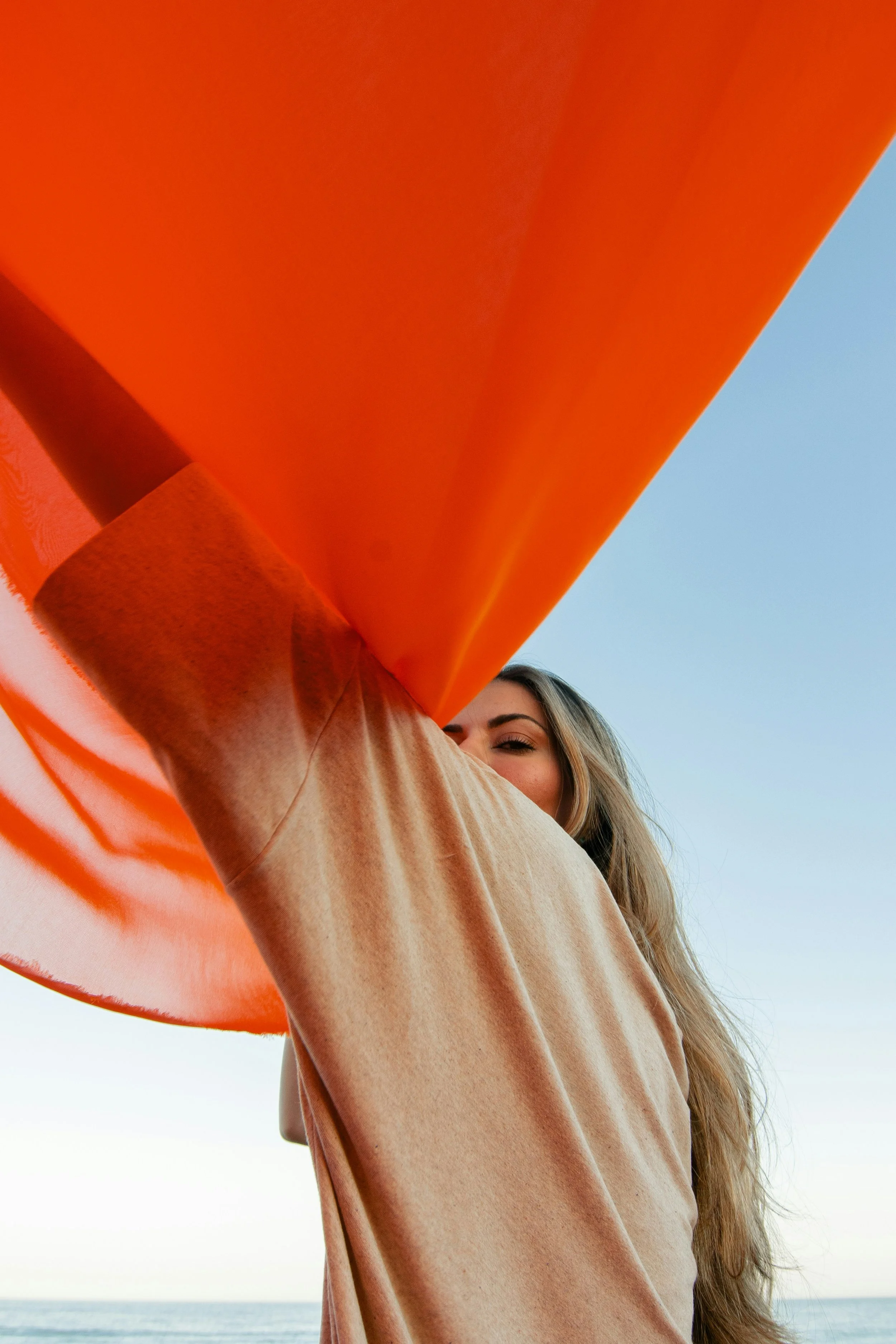 Person with long blonde hair wearing a beige sweatshirt and an orange object near their face, with a body of water and clear sky in the background.