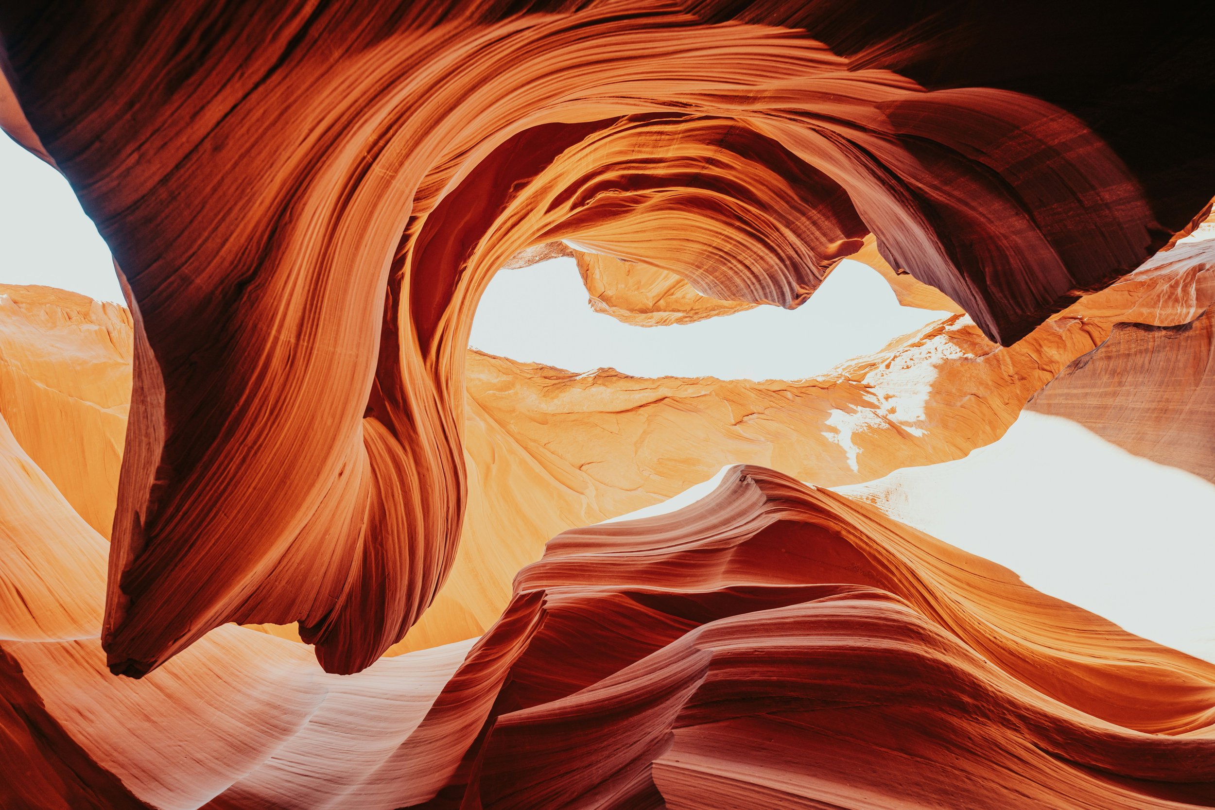 A sunlit view of carved sandstone formations with swirling patterns and vibrant orange and reddish hues in a canyon.