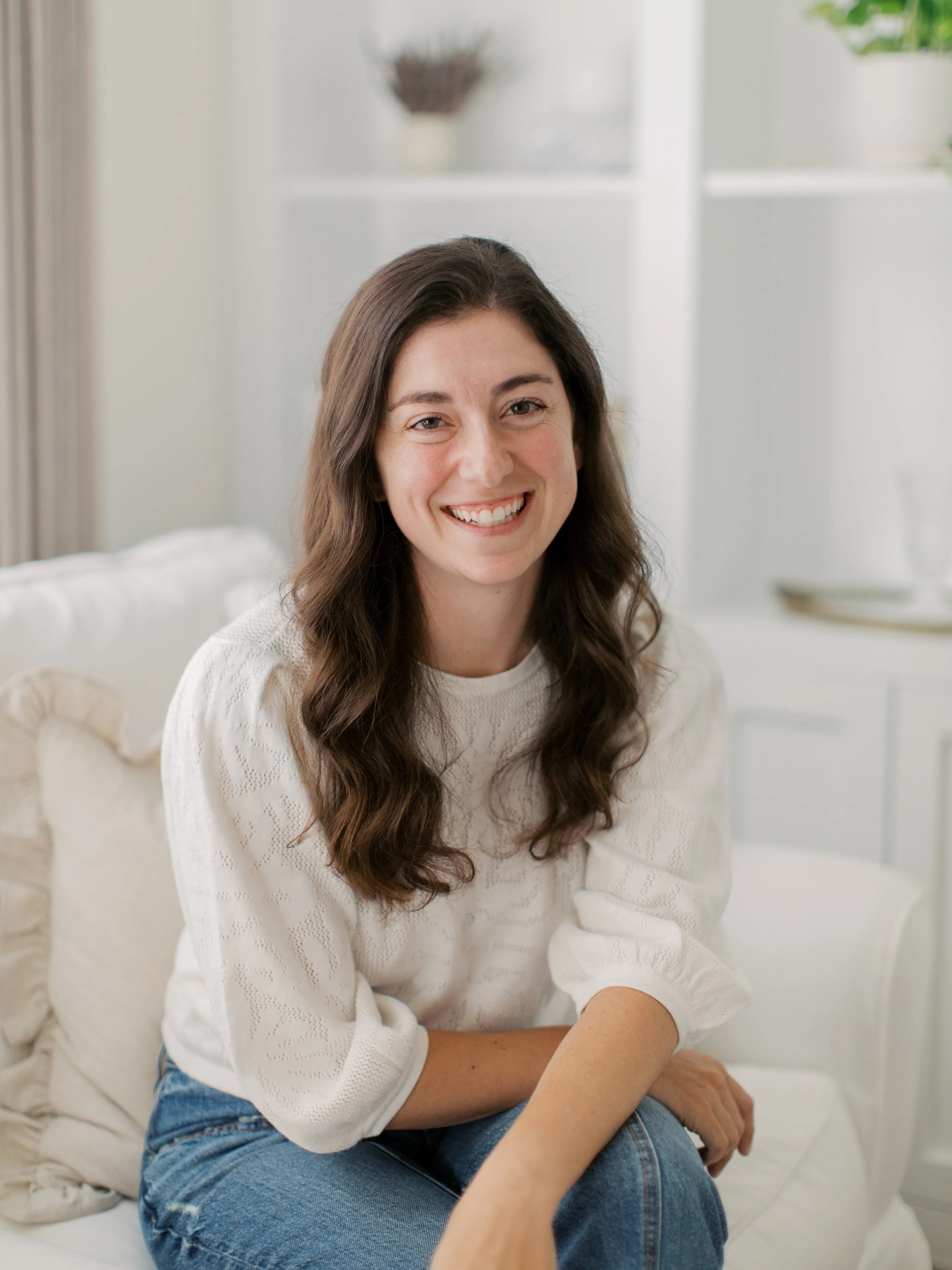 A woman with long brown hair, smiling, wearing a white sweater and blue jeans, sitting on a white bed with pillows in a bright room.