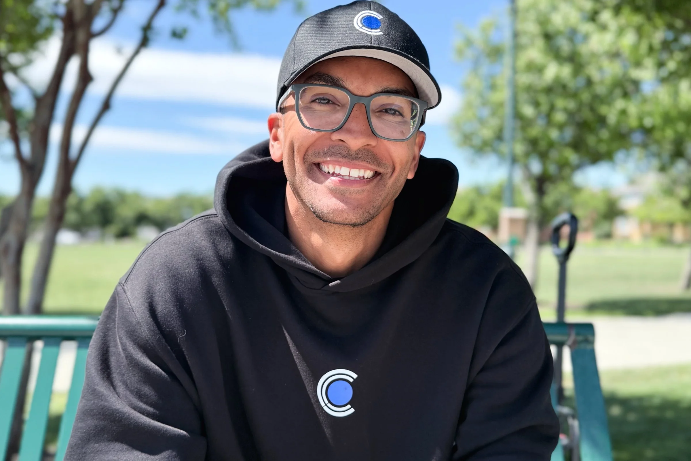 Ryan Rucker of Courtside Coaching, smiling, wearing glasses, a black hoodie, and a black baseball cap with a blue circular logo, sitting outdoors on a park bench with trees and a blue sky in the background.