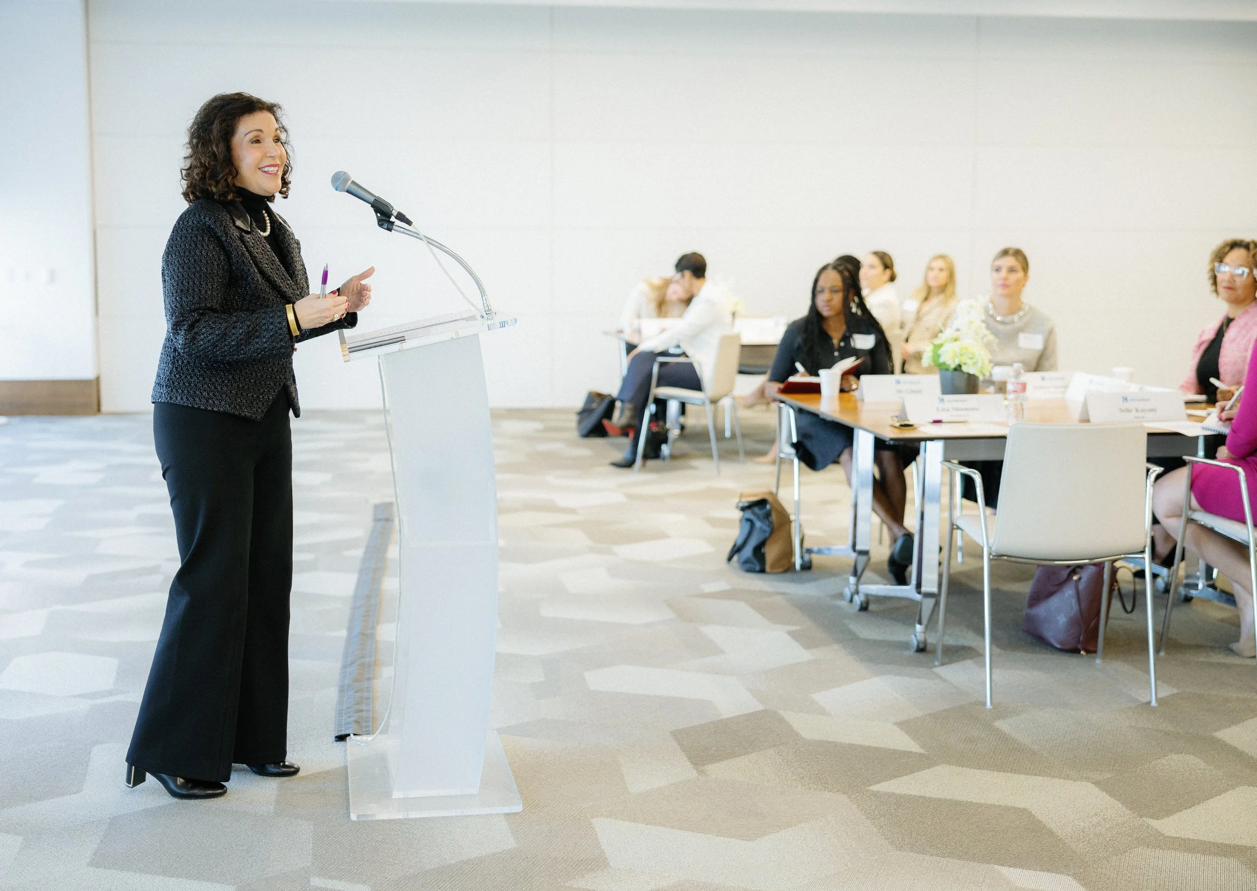 A woman in a black textured blazer and black pants standing at a clear lectern, speaking to an audience in a well-lit conference room. Several women are seated at tables, listening and taking notes.