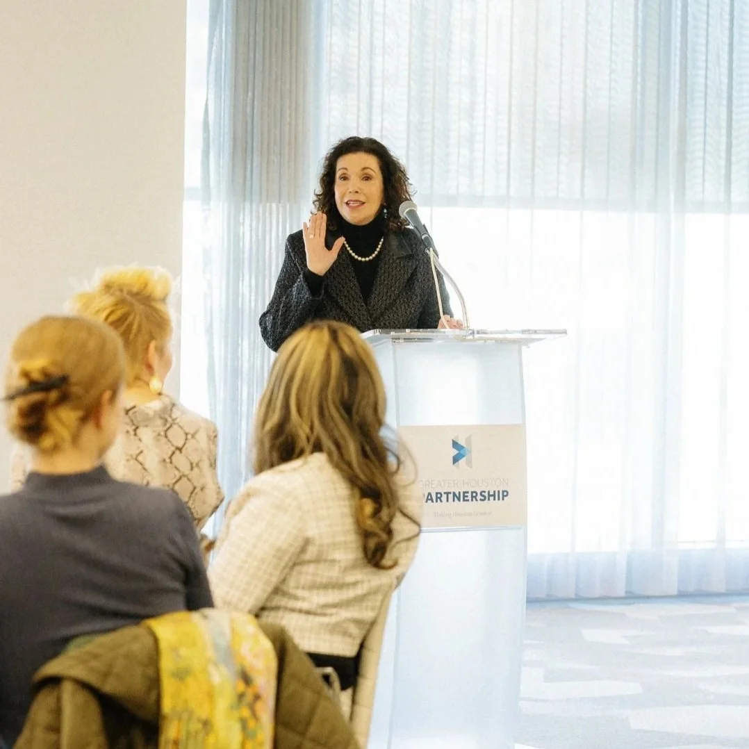 A woman speaking at a podium with a microphone at a business event, with three women seated in front listening.
