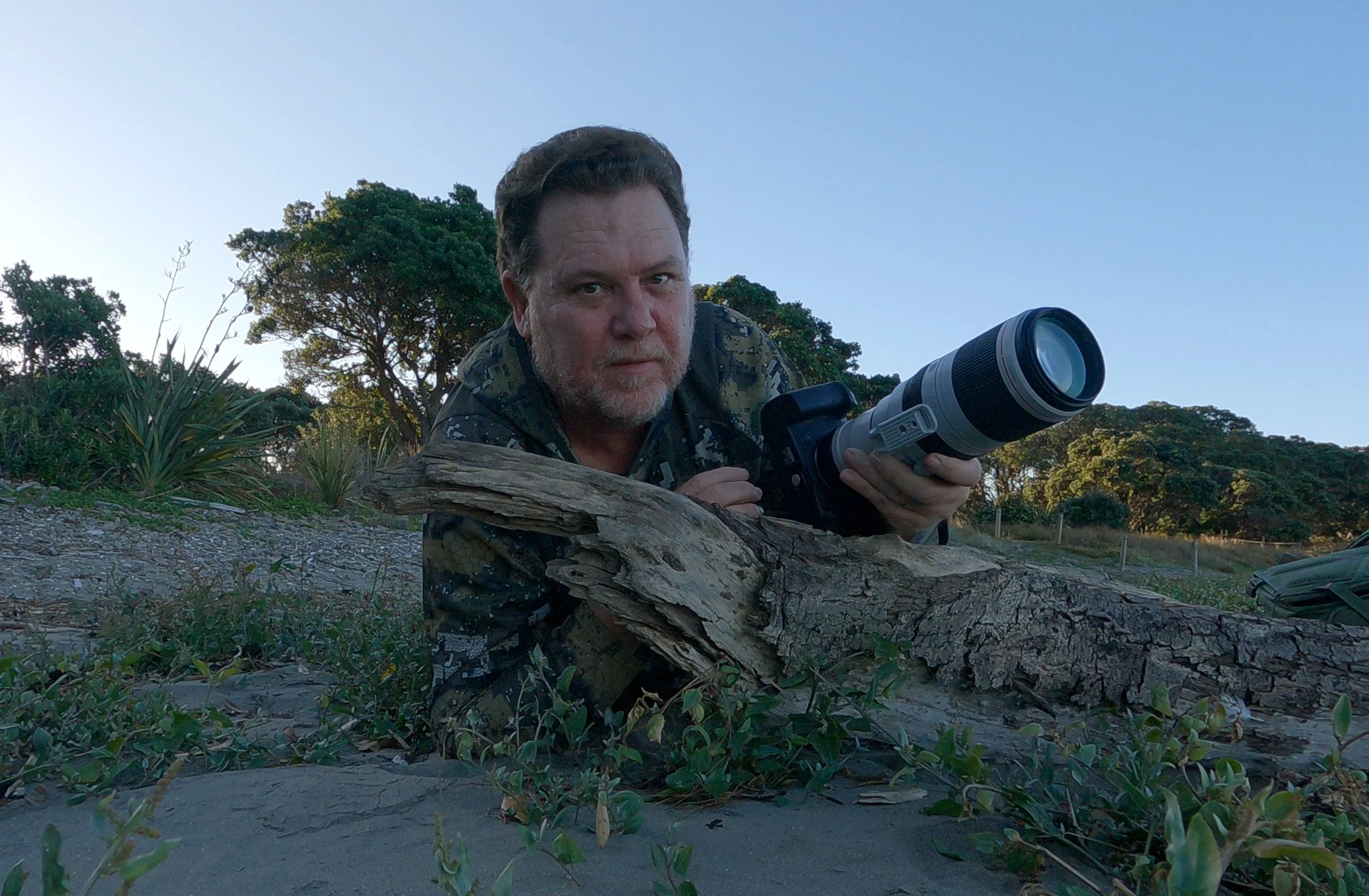 Deon, Photographer lying on the ground outdoors, holding a camera with a large telephoto lens, surrounded by low vegetation and trees under a clear blue sky.