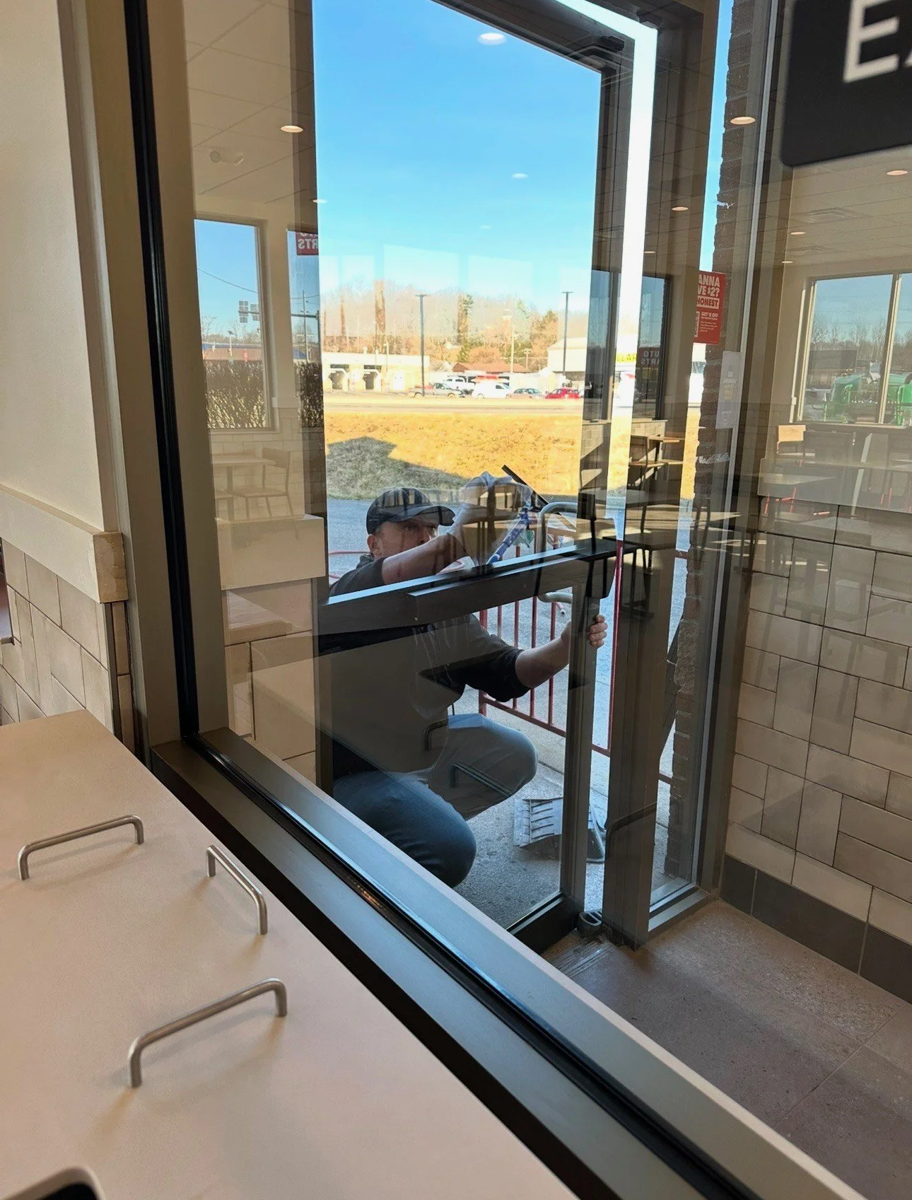 Person kneeling outside of a glass door, adjusting the door handle or lock, with the reflection of the inside of a restaurant or fast-food restaurant visible in the glass.
