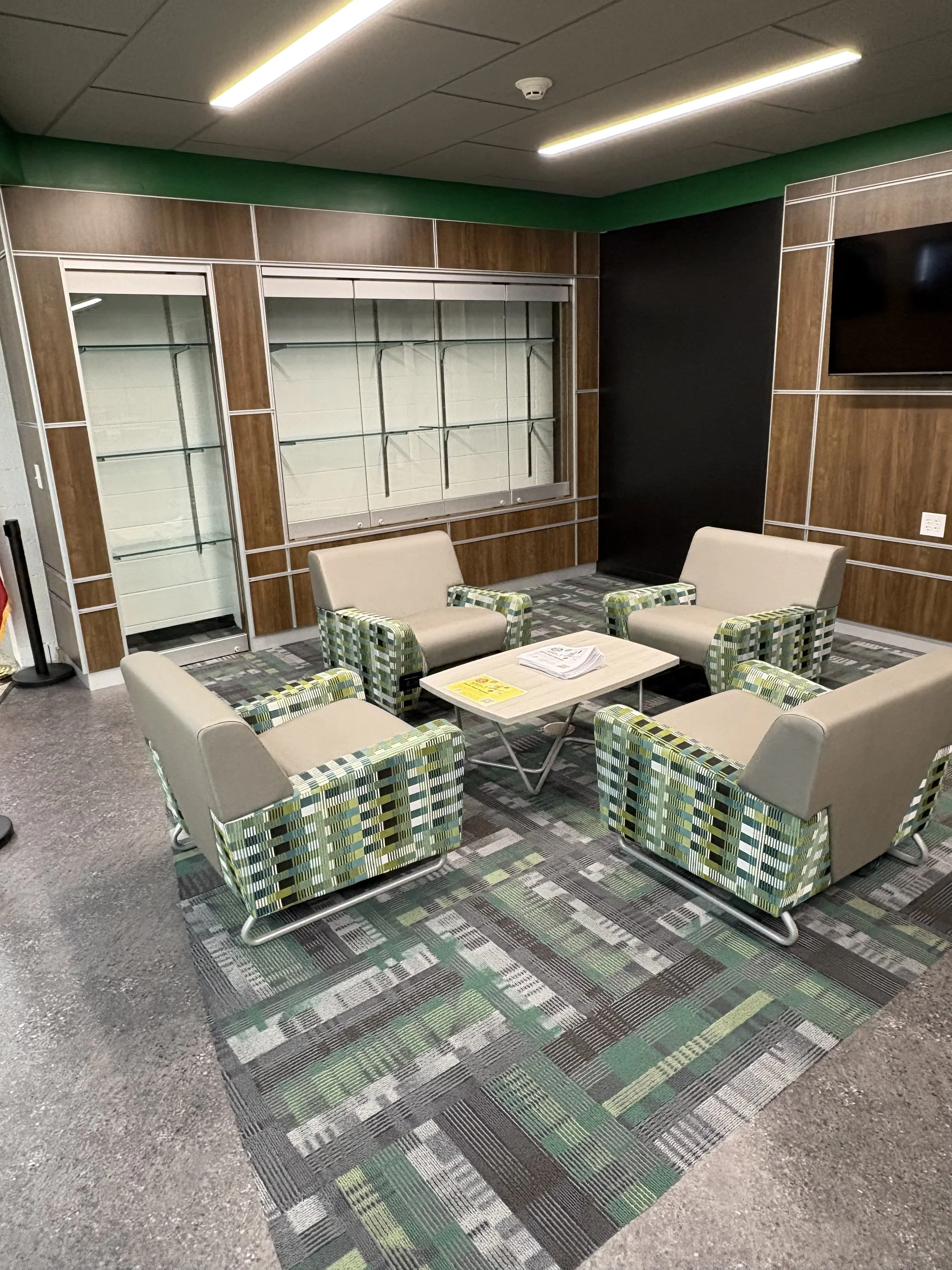 A seating area with six chairs around a small rectangular table, situated on a colorful patterned carpet. The chairs have beige cushions with patterned armrests, and the table has papers and a yellow warning sign on it. Behind the seating area, there is a wooden wall with two glass display cases and a black wall to the right, with a large mounted television screen. The ceiling has fluorescent lighting.