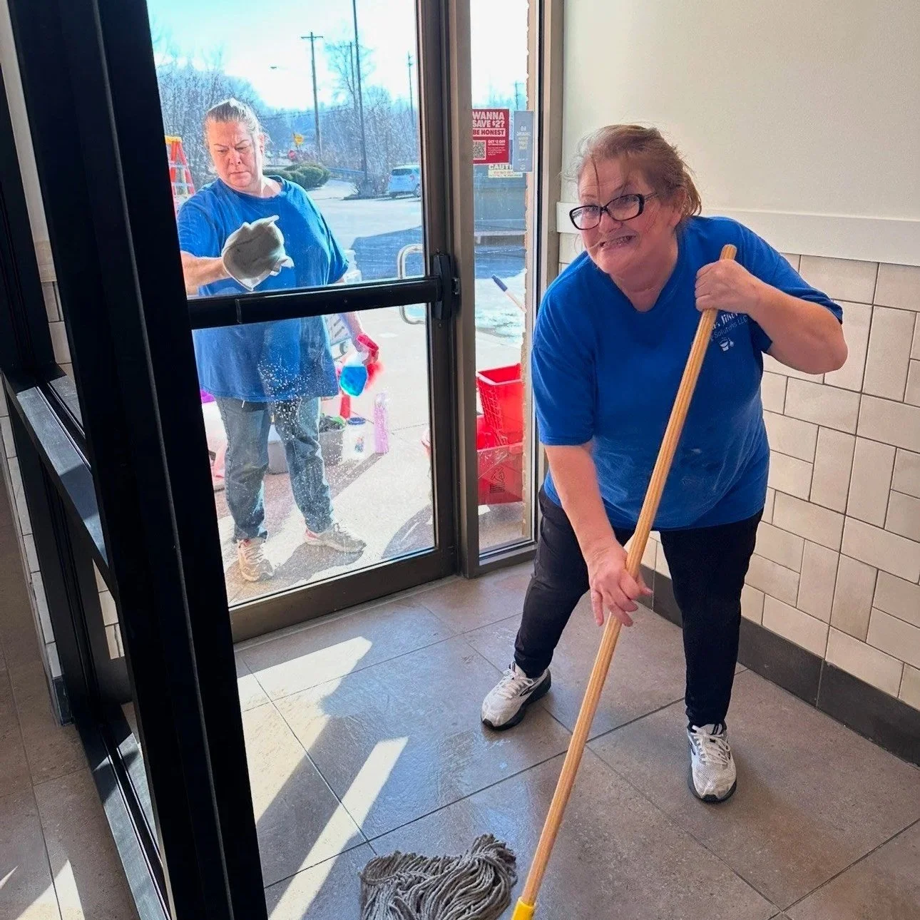 Two women cleaning the floor at a glass door entrance. One woman is inside cleaning the floor with a mop, and the other woman is outside cleaning the glass door with a cloth.