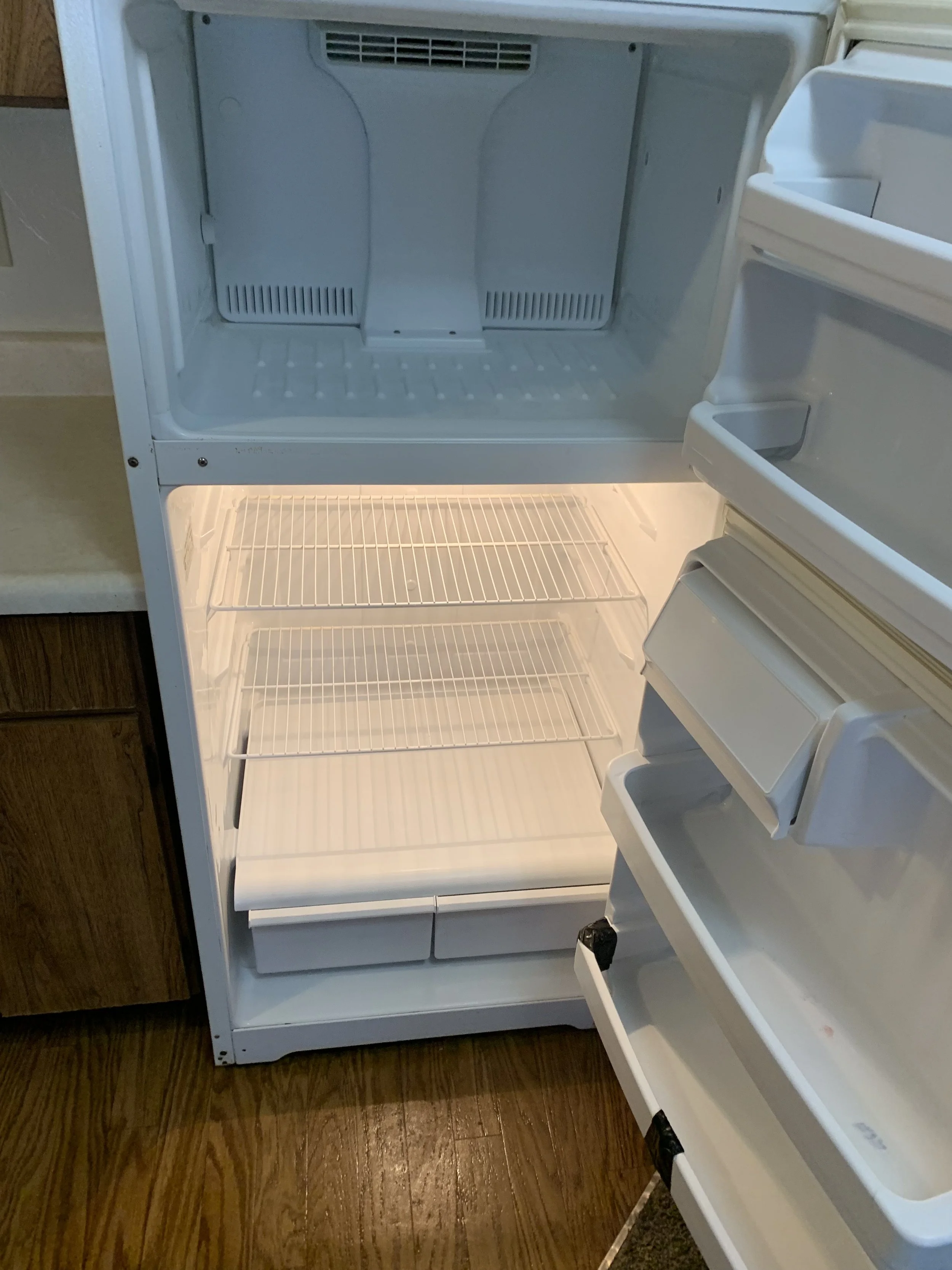 Empty white refrigerator with shelves, drawers, and door compartments, open and illuminated, on a wooden floor in a kitchen.