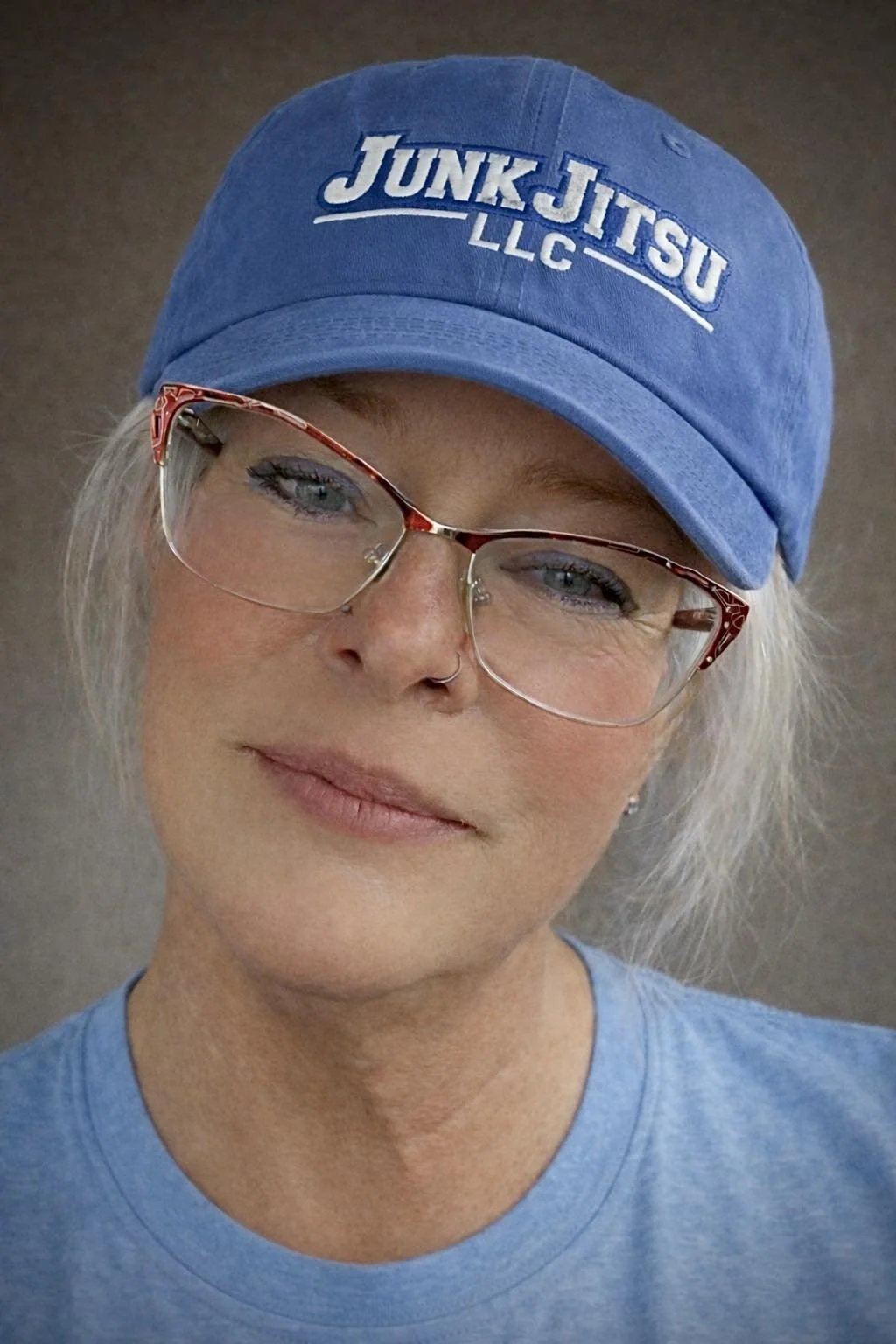 A woman wearing glasses, a blue cap with the words 'Junk Jitsu LLC', and a matching blue shirt, smiling softly.