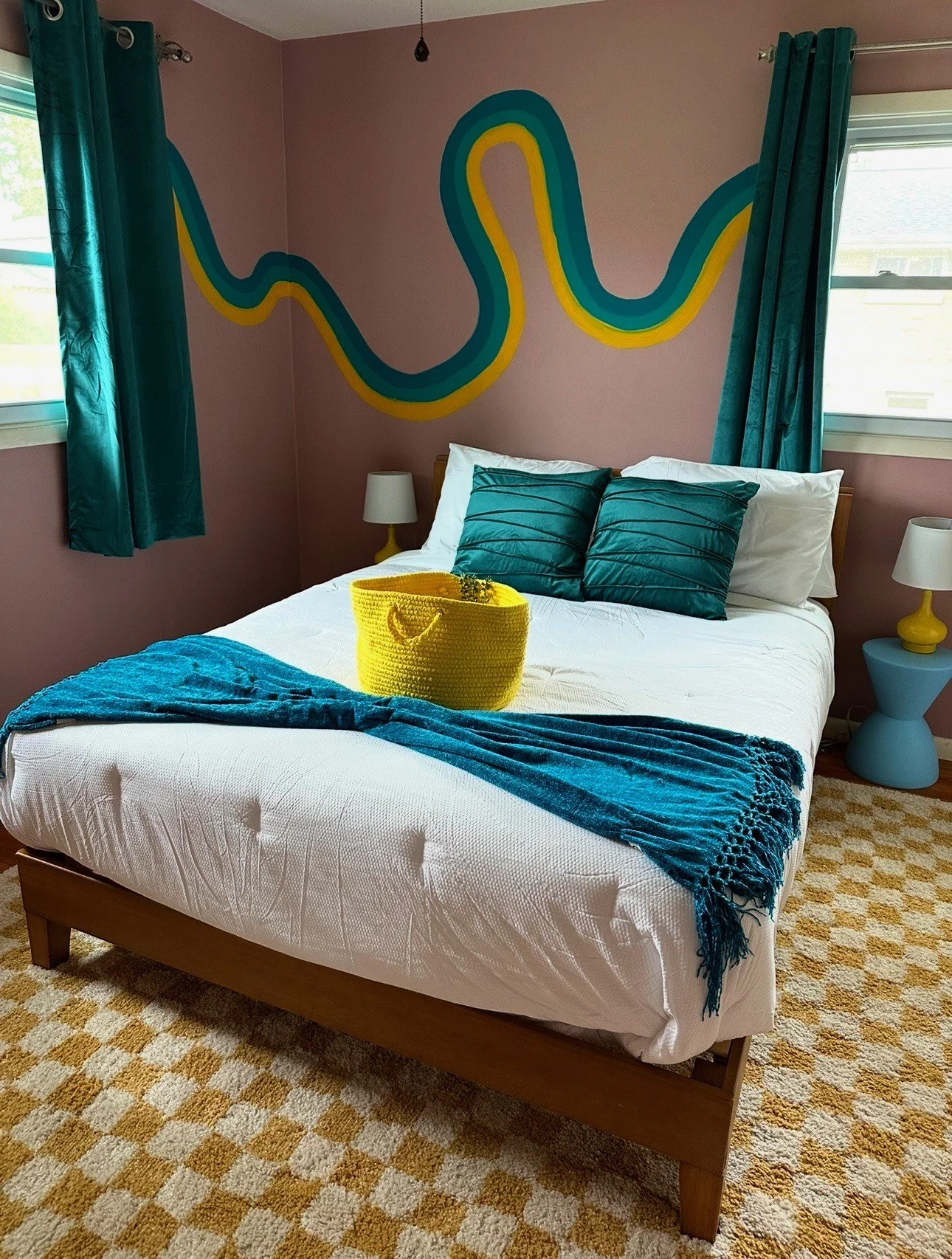 A bedroom with pink walls, featuring a colorful wavy mural above the bed. The bed has white bedding, two teal pillows, a blue throw blanket, and a yellow knitted basket on top. There are teal curtains on both windows, and matching lamps on blue bedside tables.