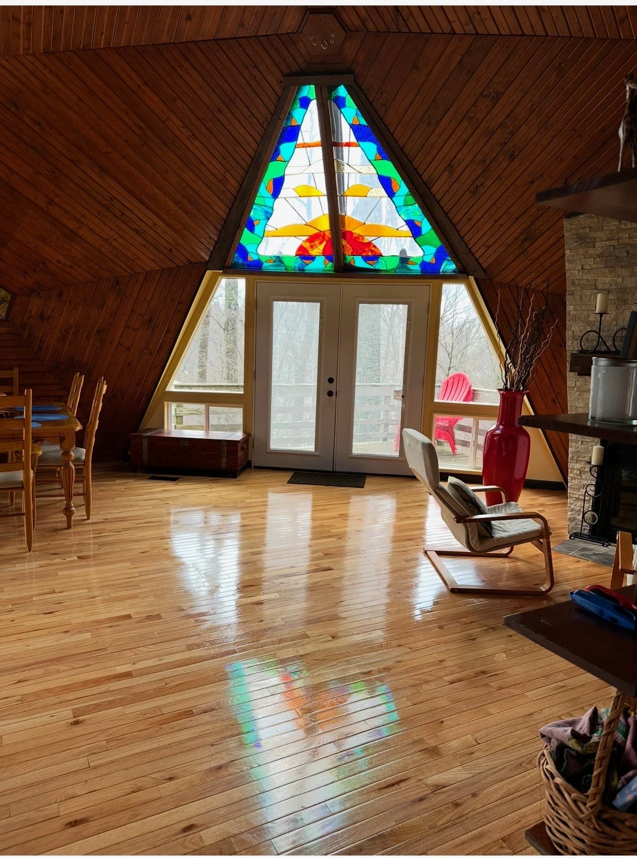 Interior view of a room with glossy hardwood floors, wooden walls, a large stained glass window with a colorful sun and sky design, glass doors leading to a balcony with red chairs, a wooden dining table with chairs, a white armchair, a large red vase with branches, and a stone fireplace.