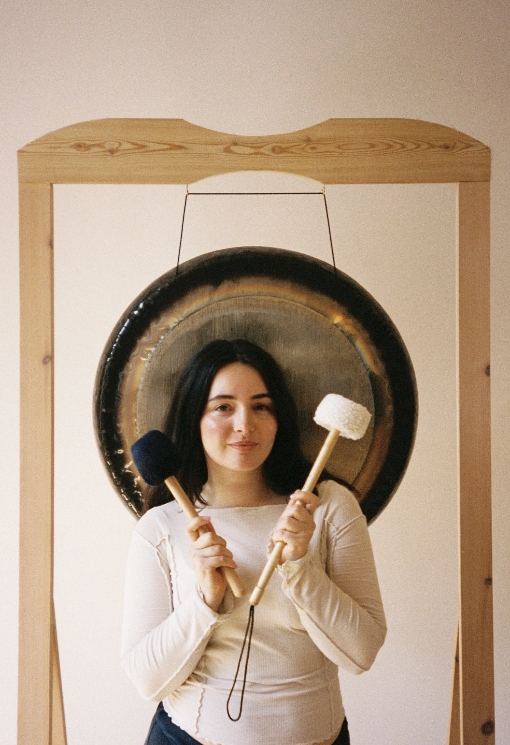 A woman with dark hair holding two mallets standing in front of a large gong mounted on a wooden frame.