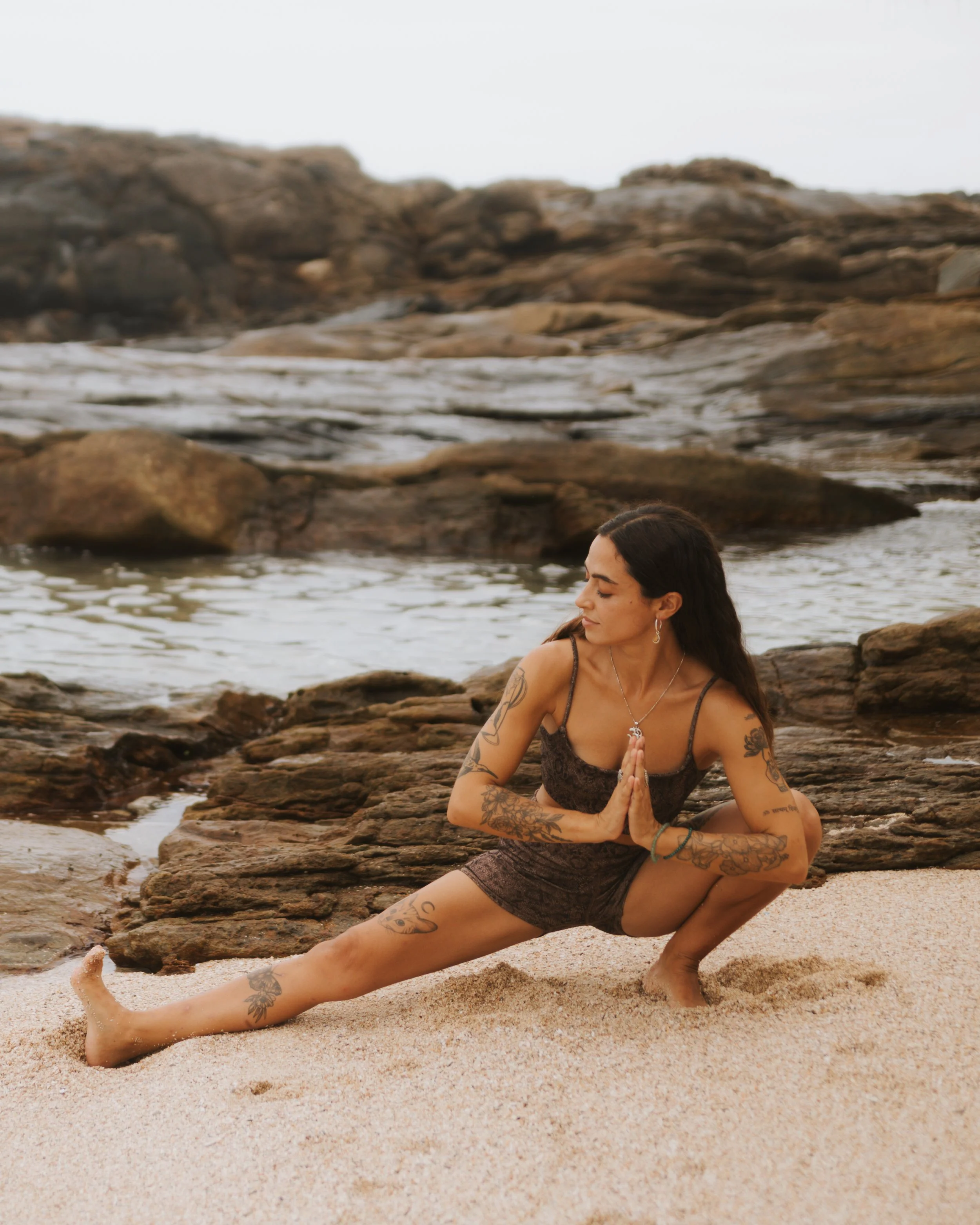 A woman practicing yoga on a sandy beach near rocks and water, wearing a dark outfit and tattoos, in a lunge pose with hands in prayer position.