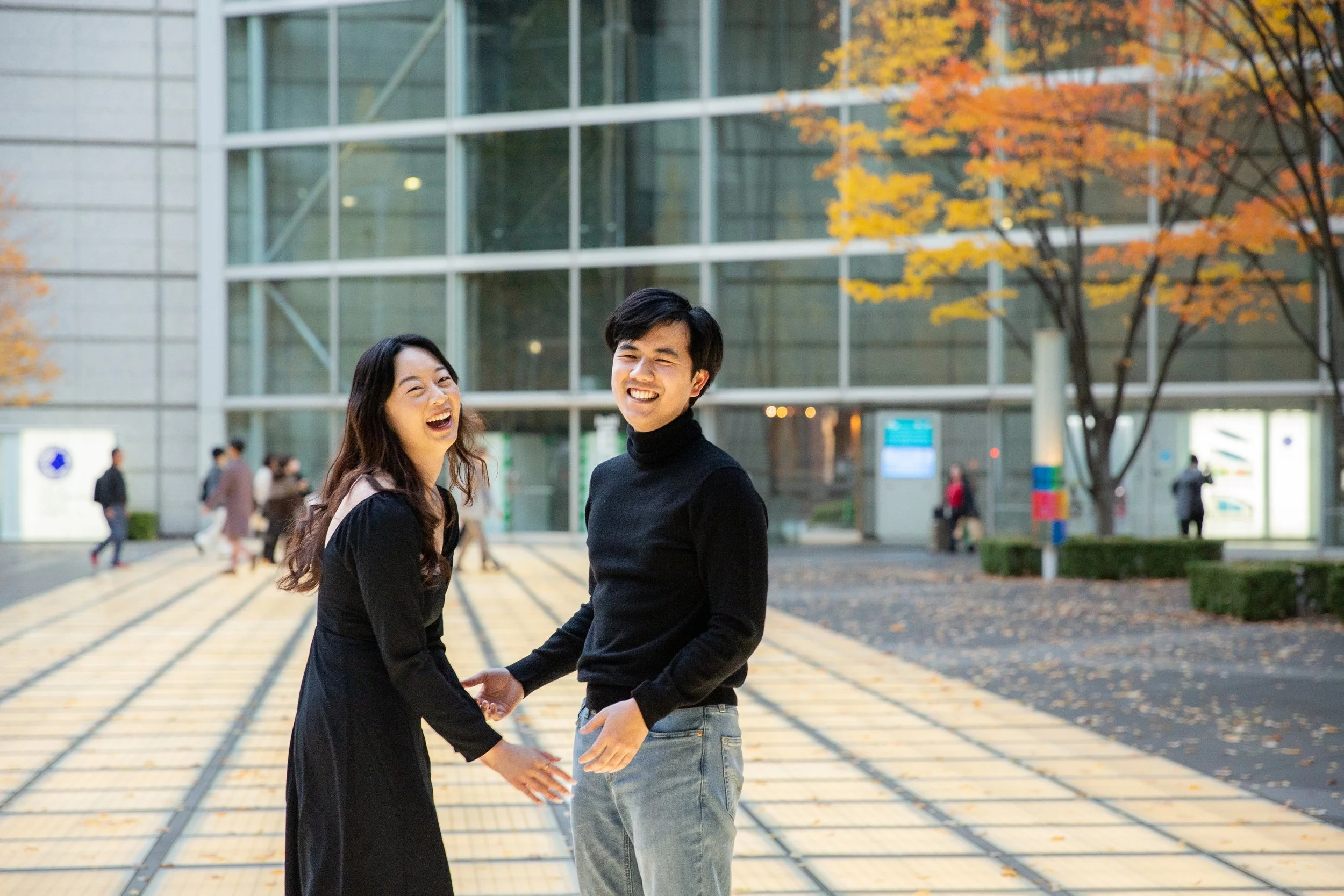 Tokyo Portrait Photographer, couple in Tokyo, fall, modern building, 笑顔の若いカップルが手を取り合っている。背景には秋の落ち葉が散る樹木と現代的なビルの建物。
