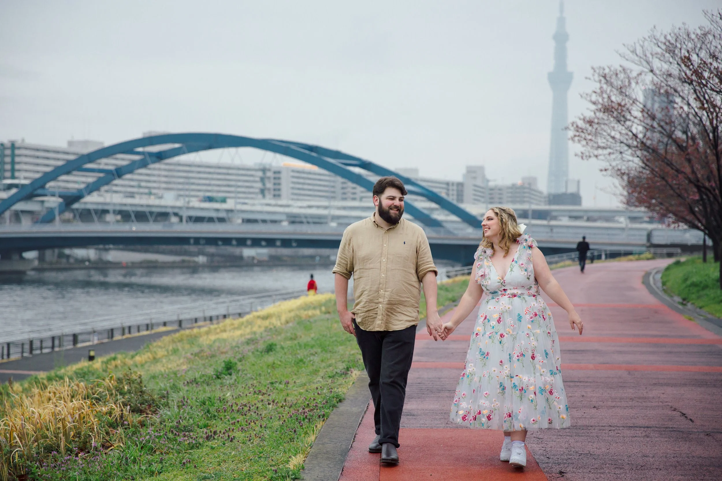 Tokyo Portrait Photographer, Sky Tree, riverside, bridge in Tokyo, couple holding hands, 川沿いの遊歩道を散歩する若いカップル。背景に都市のビルと東京スカイツリーが見える。女性は花柄のワンピースを着て、男性はカジュアルなシャツを着ている。
