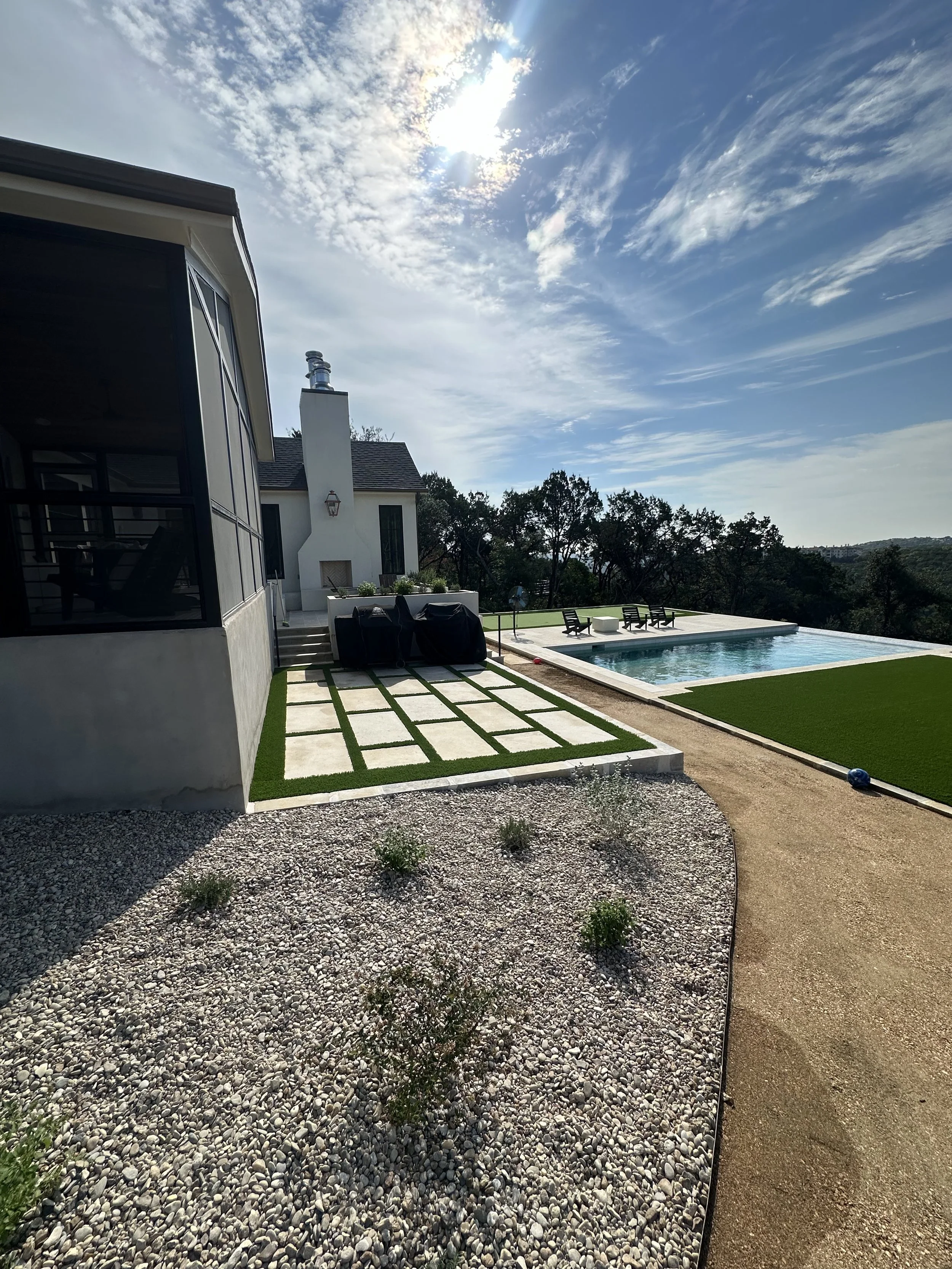 Modern backyard landscape design by Golden View Landscape Design in Austin, featuring large stone pavers with artificial turf ribbons, a limestone gravel garden, and a custom swimming pool with a scenic hill country view.