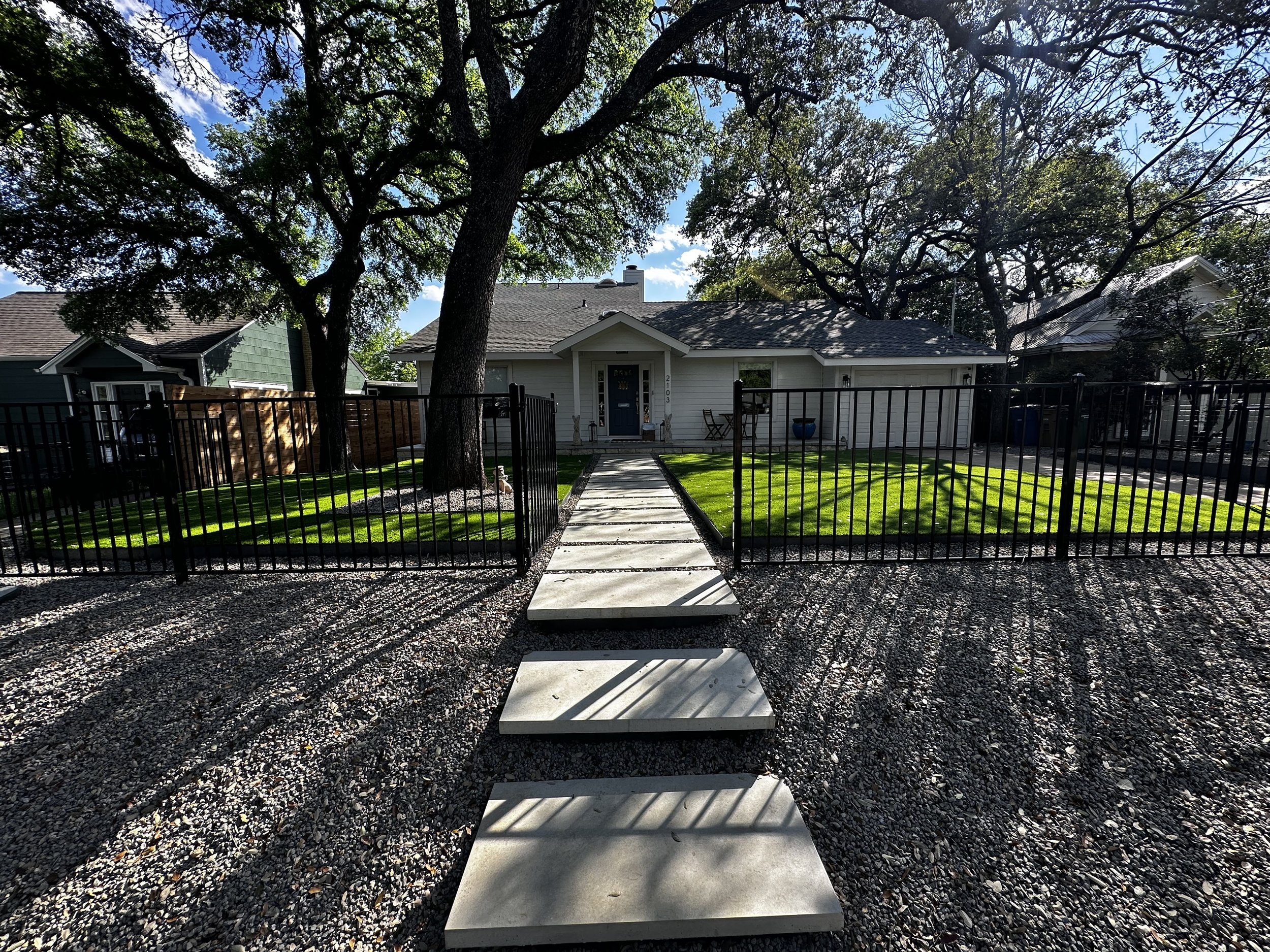 Front yard of a house with a concrete pathway, a grassy lawn, a large tree, and a black metal fence in a suburban neighborhood.