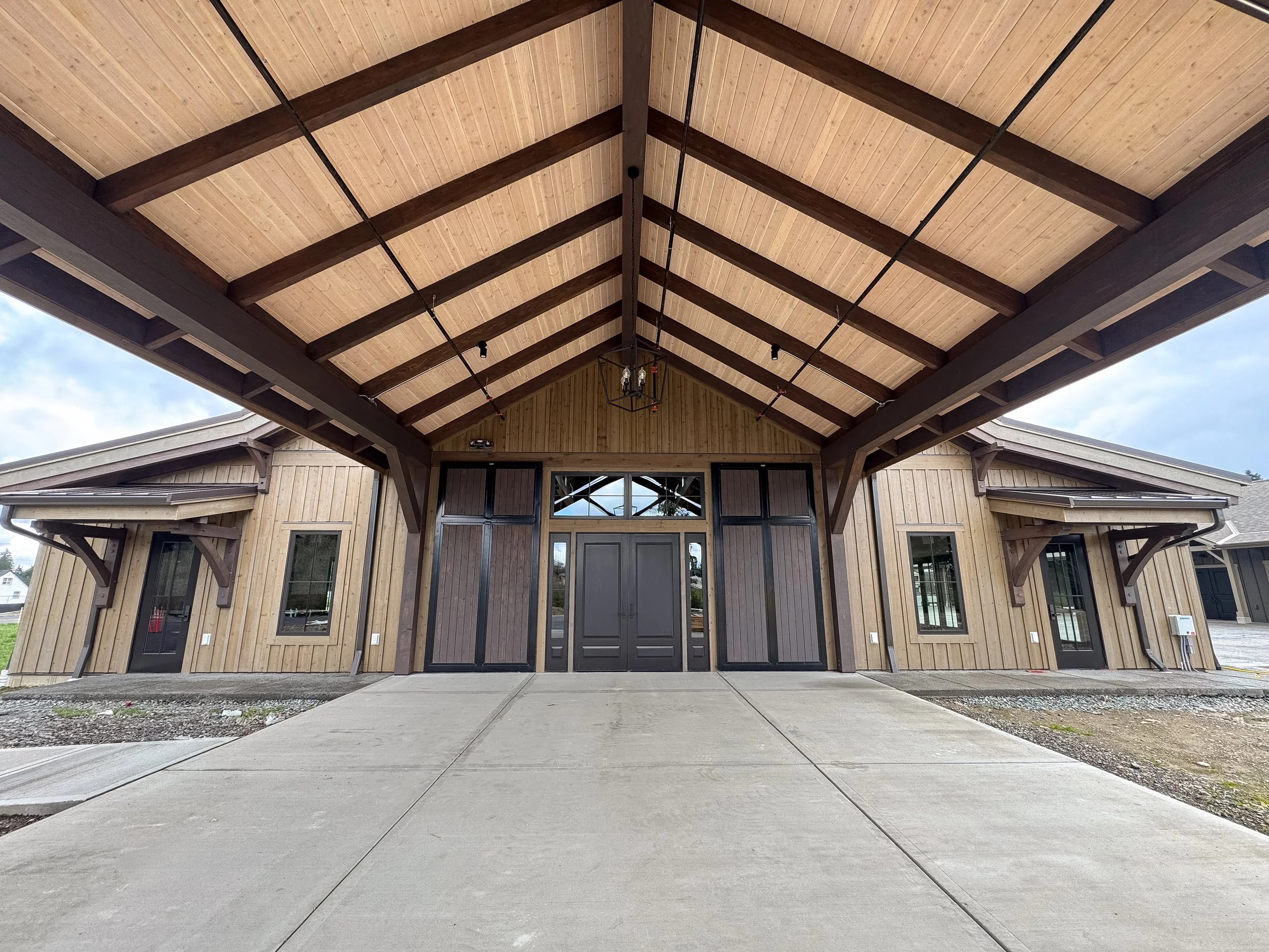 Under construction building with wooden exterior, large front entrance, and a covered porch, construction equipment and gravel in the foreground.