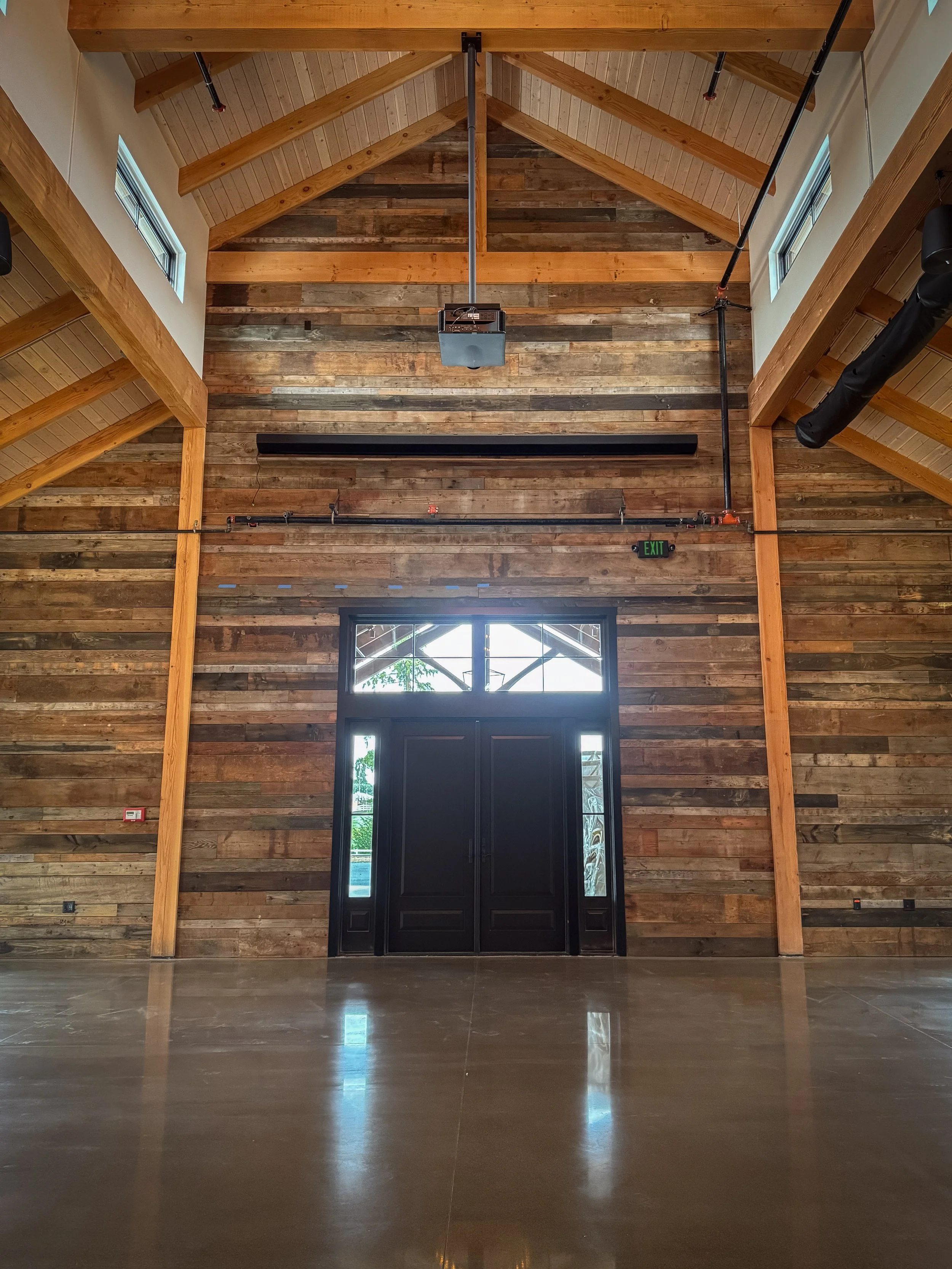 Interior of a large building under construction with exposed wooden roof beams, multiple windows, and construction equipment on the unfinished floor.