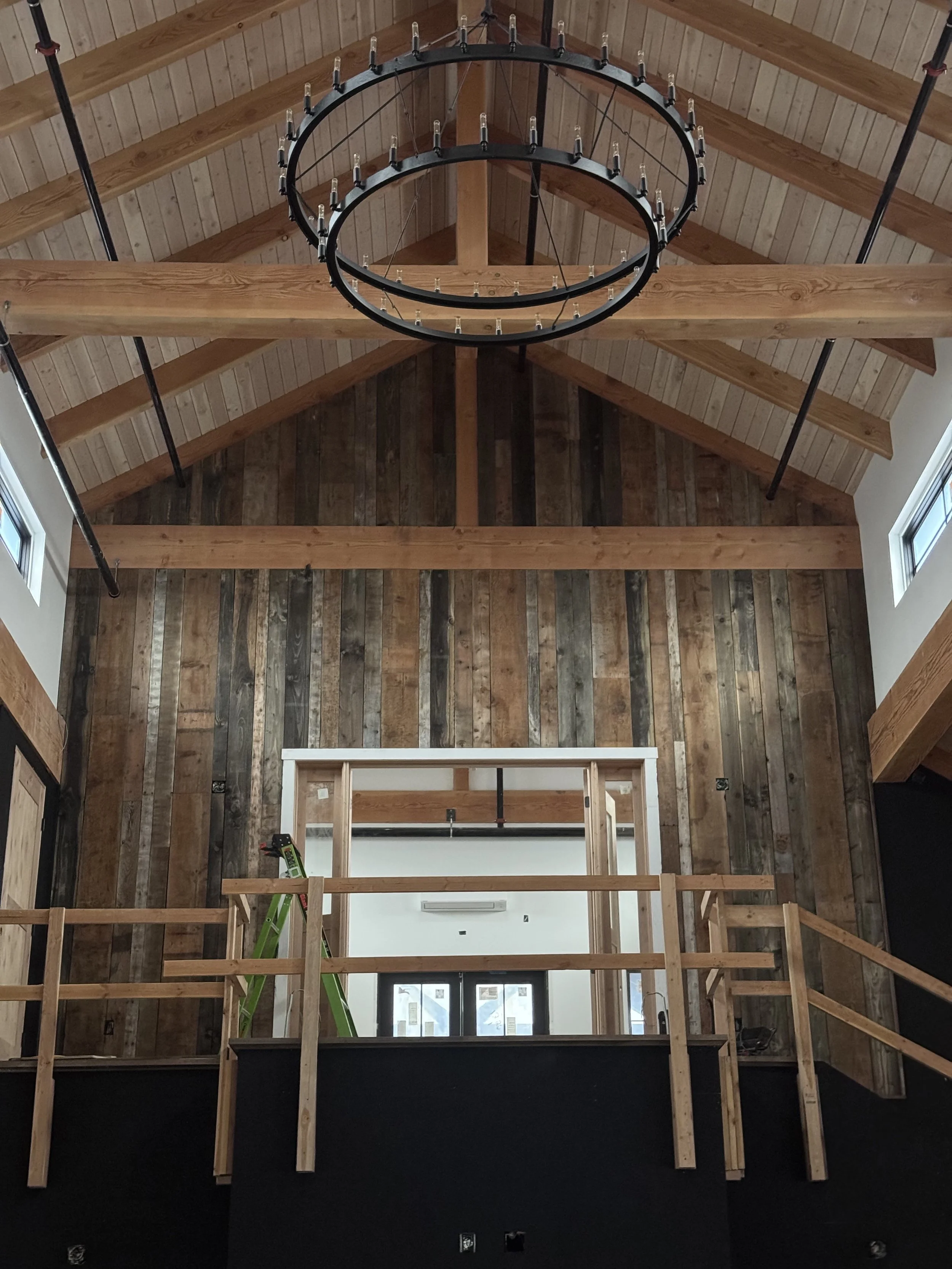 Interior of a room under construction with a high wooden ceiling, a large circular chandelier, and a partial wooden railing.