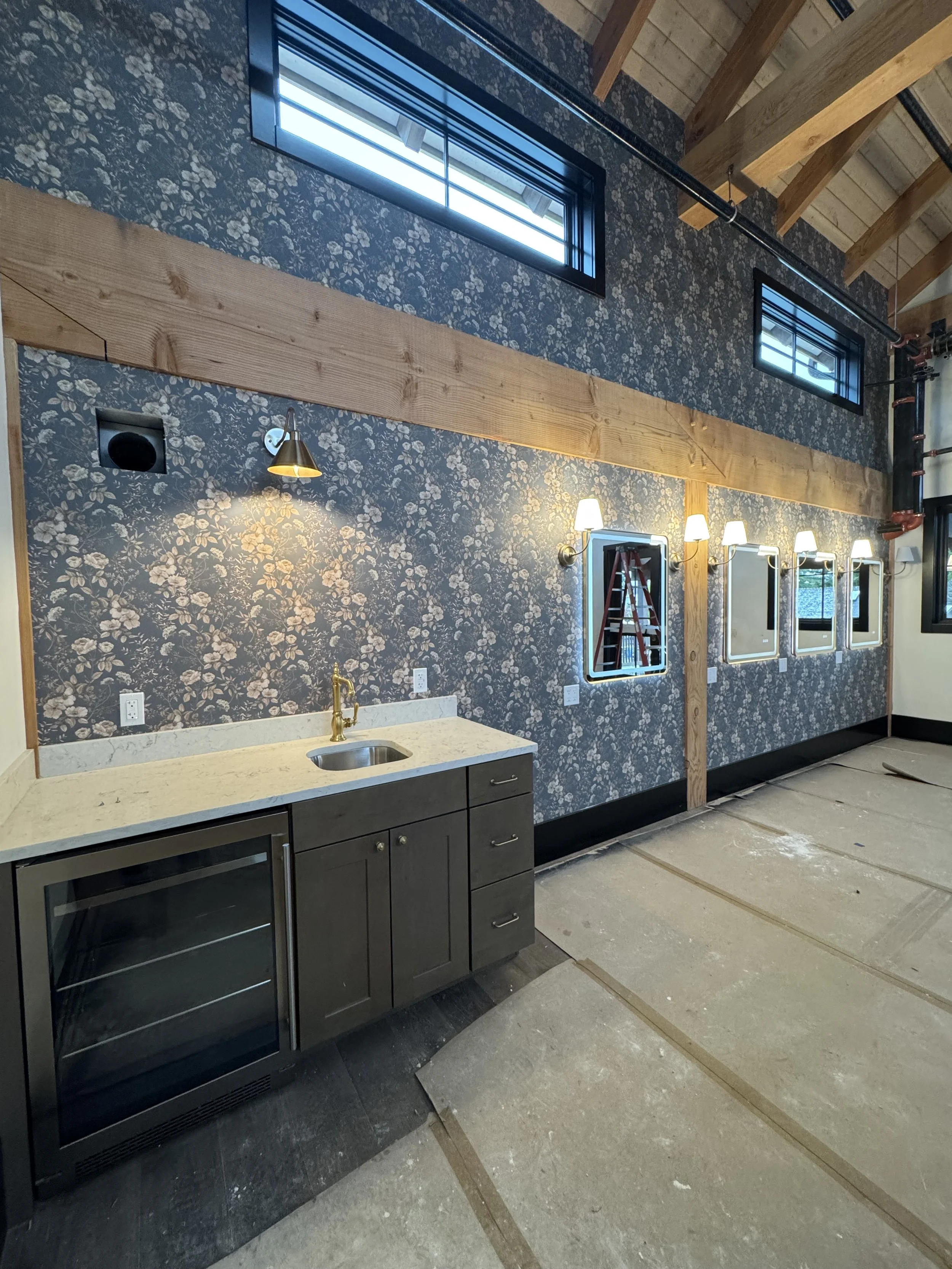 Interior of a room under construction with unfinished floors, white walls, exposed wooden beams, and framed openings for windows or doors, scaffolding in the center, and construction tools and materials scattered on the floor.