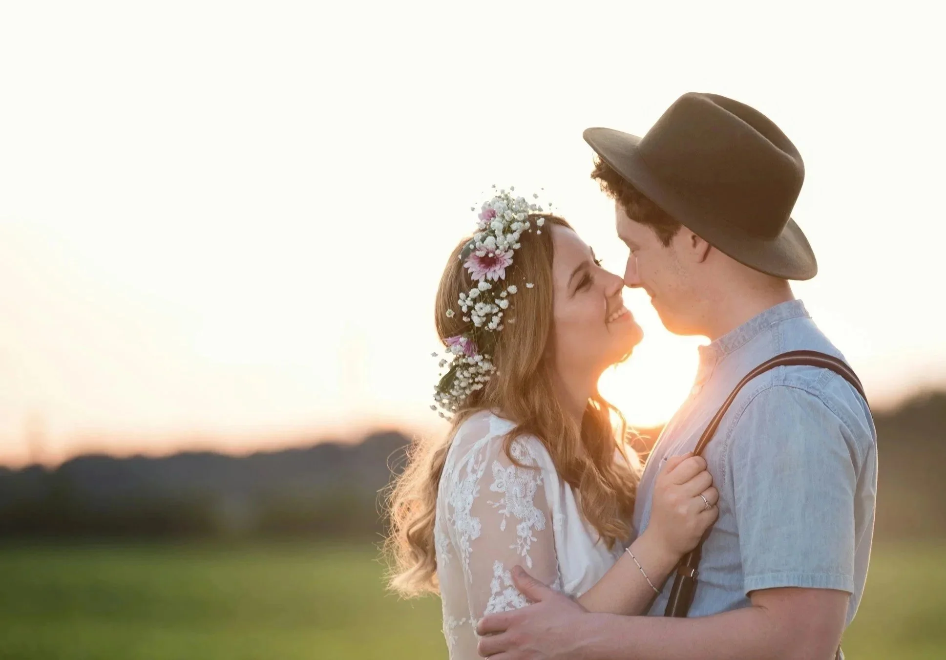 a young couple standing close together at sunset, smiling, with their noses touching. The woman is wearing a floral crown and a white lace dress, while the man is wearing a hat and a light blue shirt.