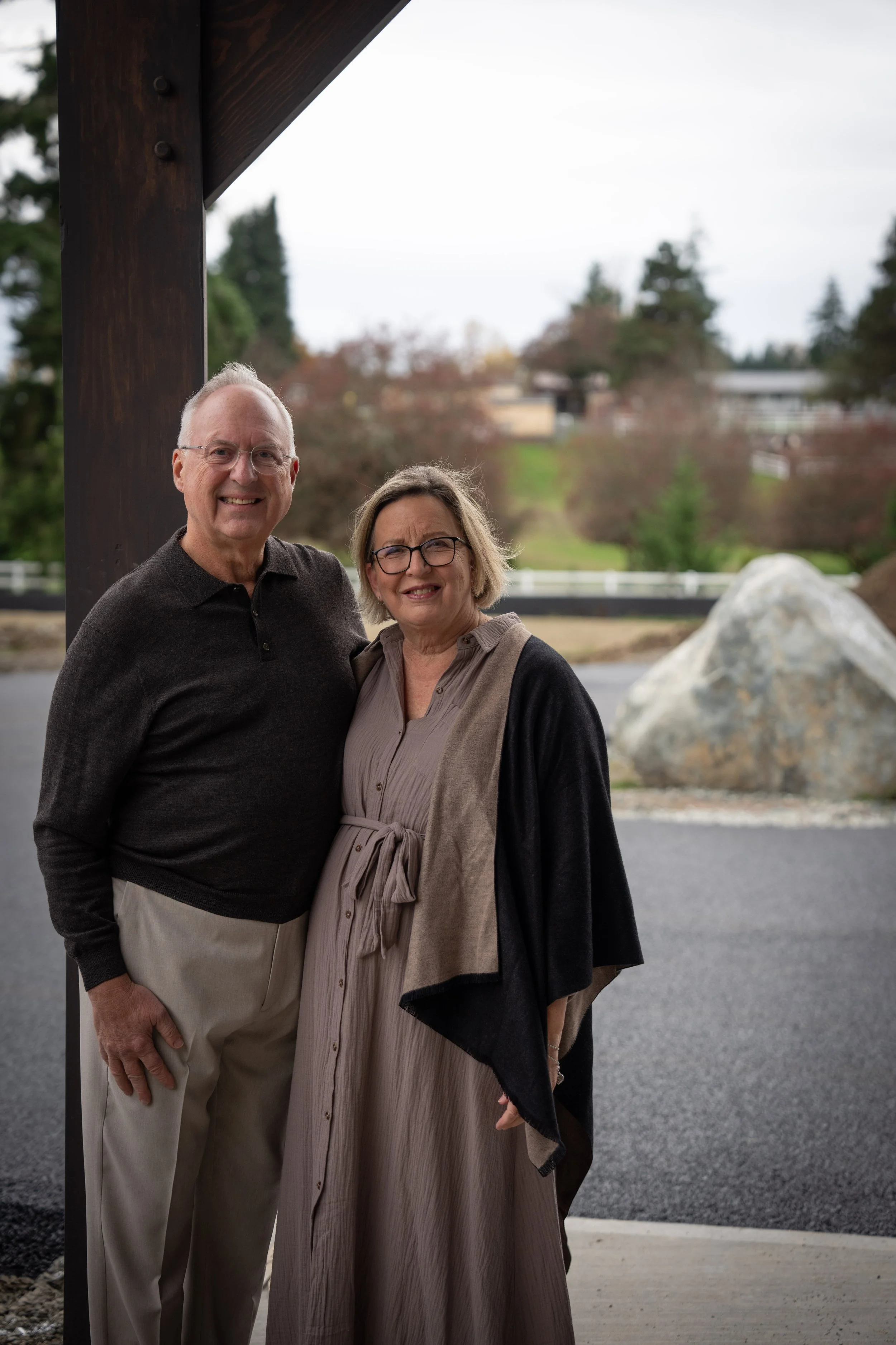 Joy & Glory Farm owners Gordon and Patty Rody standing near their beautiful new event venue, smiling at the camera, with trees, a large rock, and a road in the background.