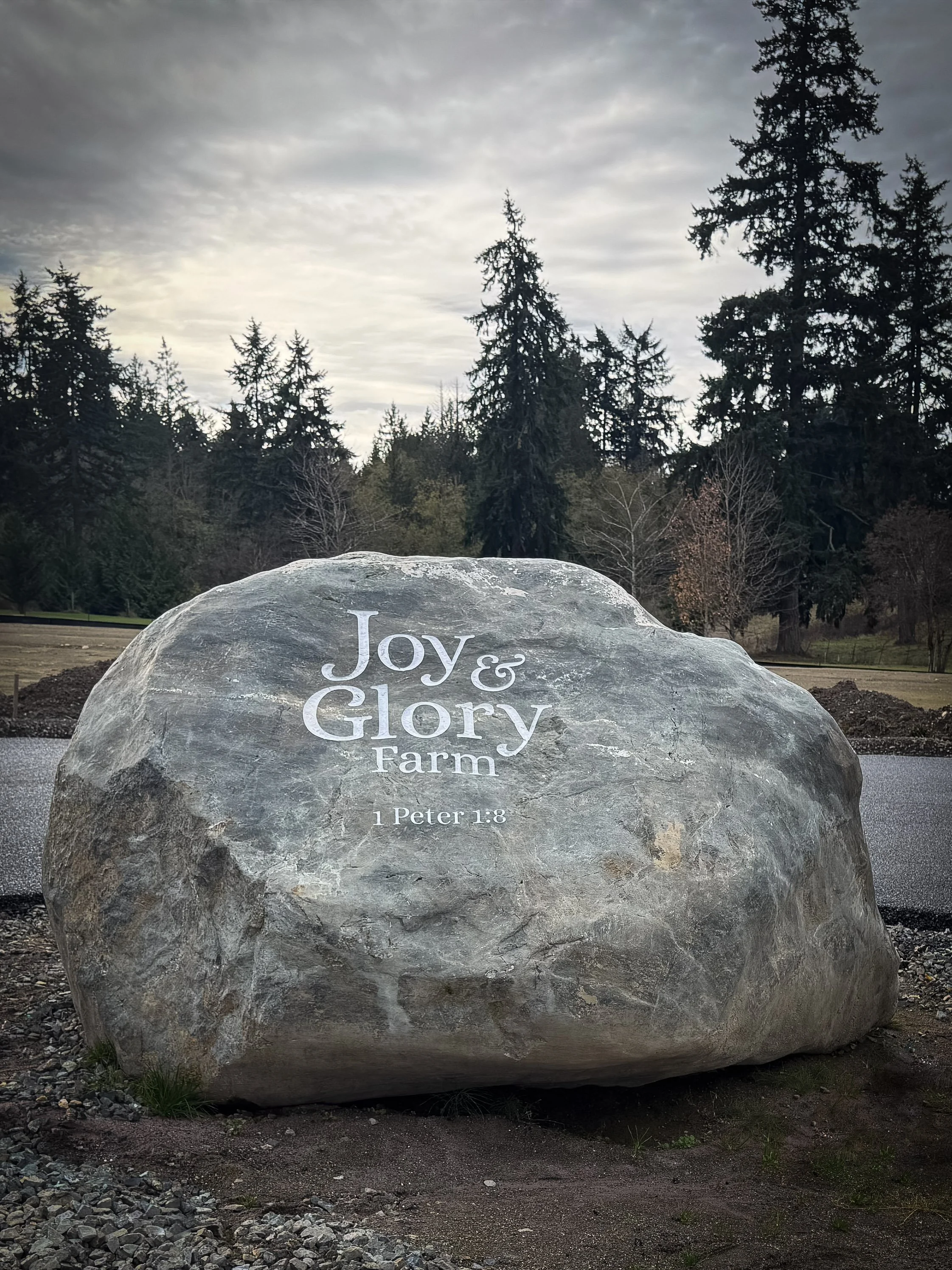 Large stone with inscription that reads "Joy & Glory Farm" and Bible reference "1 Peter 1:8." Trees in fall colors and cloudy sky in the background.