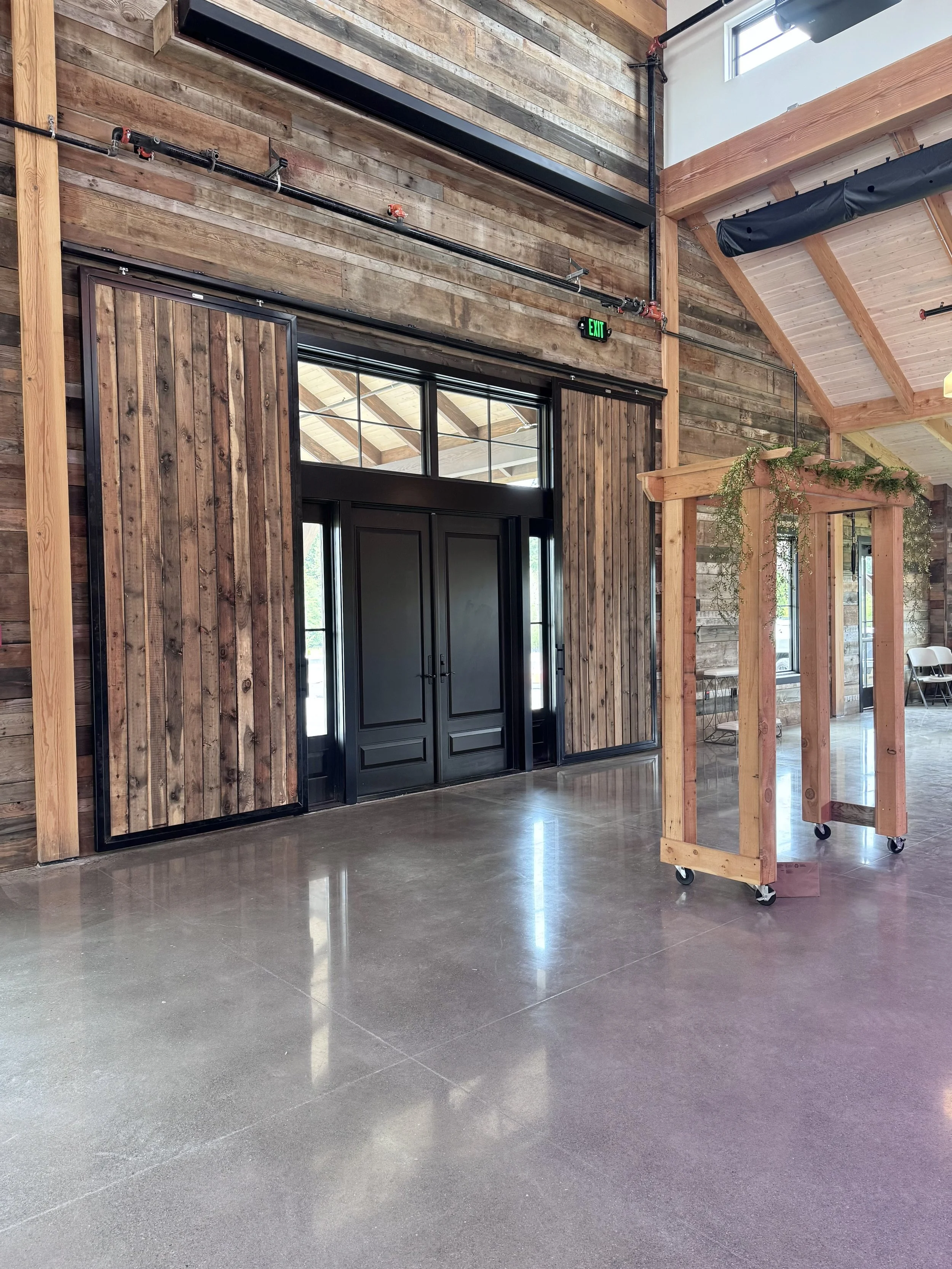 Interior of a large building under construction with exposed wooden roof beams, multiple windows, and construction equipment on the unfinished floor.