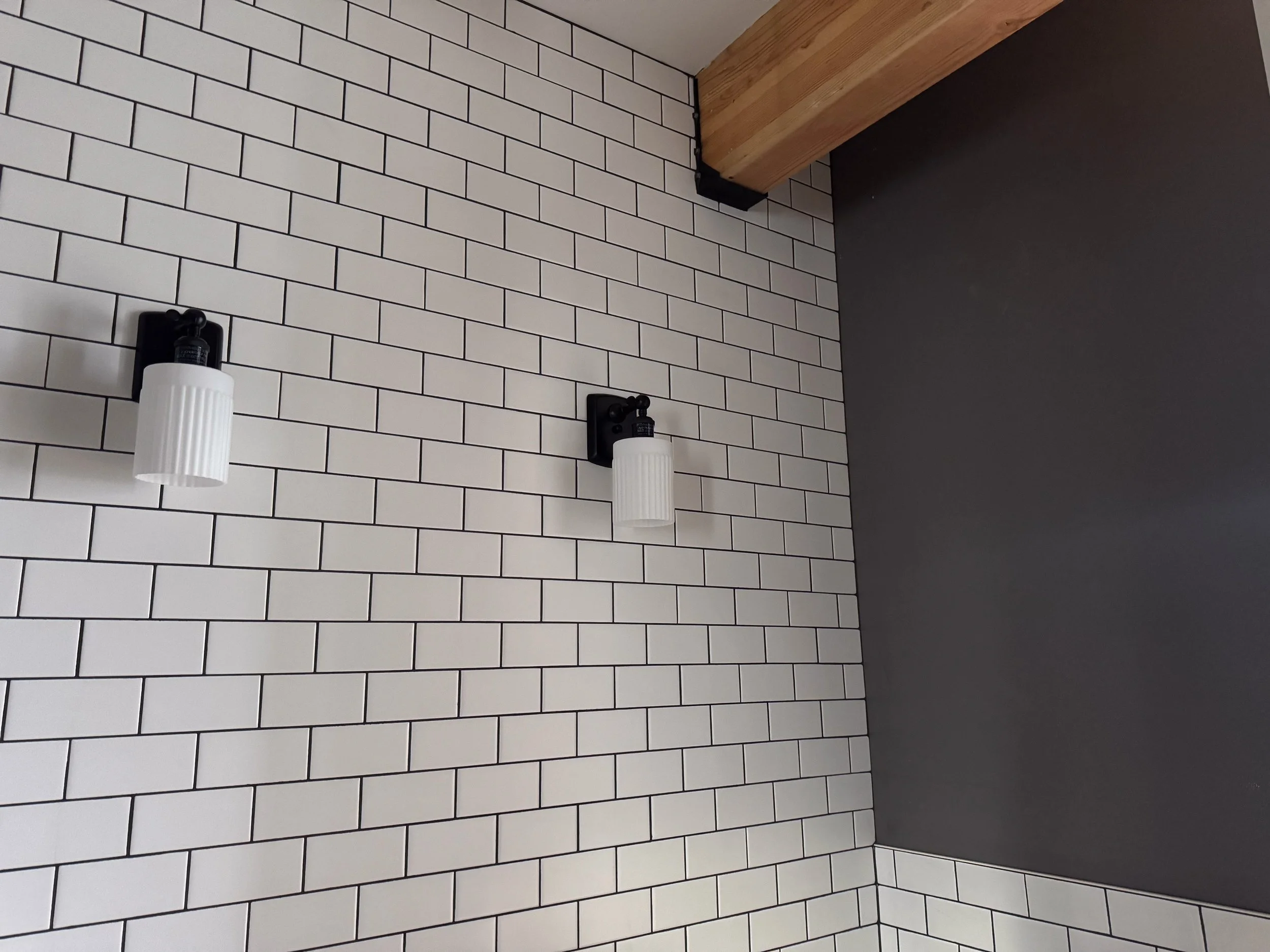 Two modern wall-mounted light fixtures with ribbed white glass shades on a white subway tiled wall in a room with a grey painted wall and wooden ceiling beam.