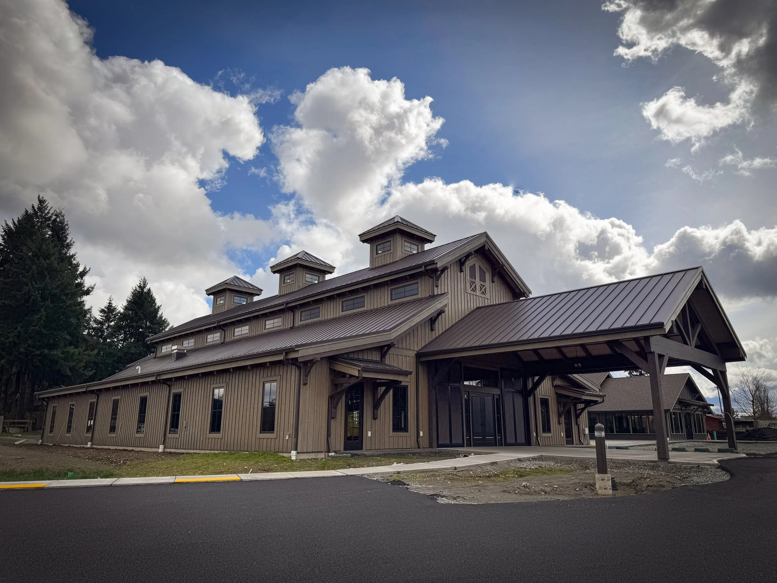 Construction site of a large wooden building with a metal roof, surrounded by construction equipment and barriers, under a cloudy sky.