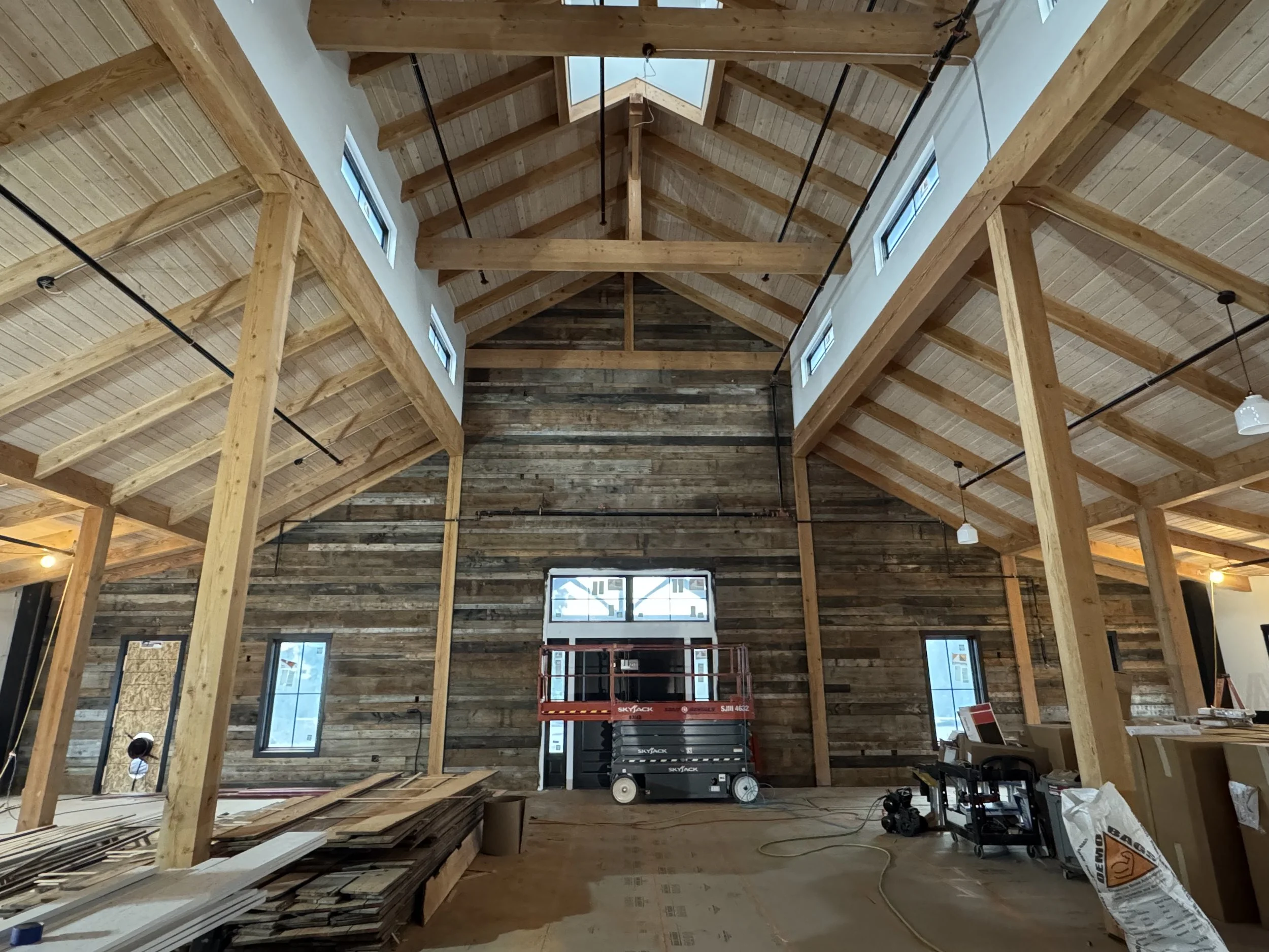 Interior of a building under construction with a high wooden ceiling, exposed beams, and a wooden wall. Construction equipment and materials are scattered on the floor.