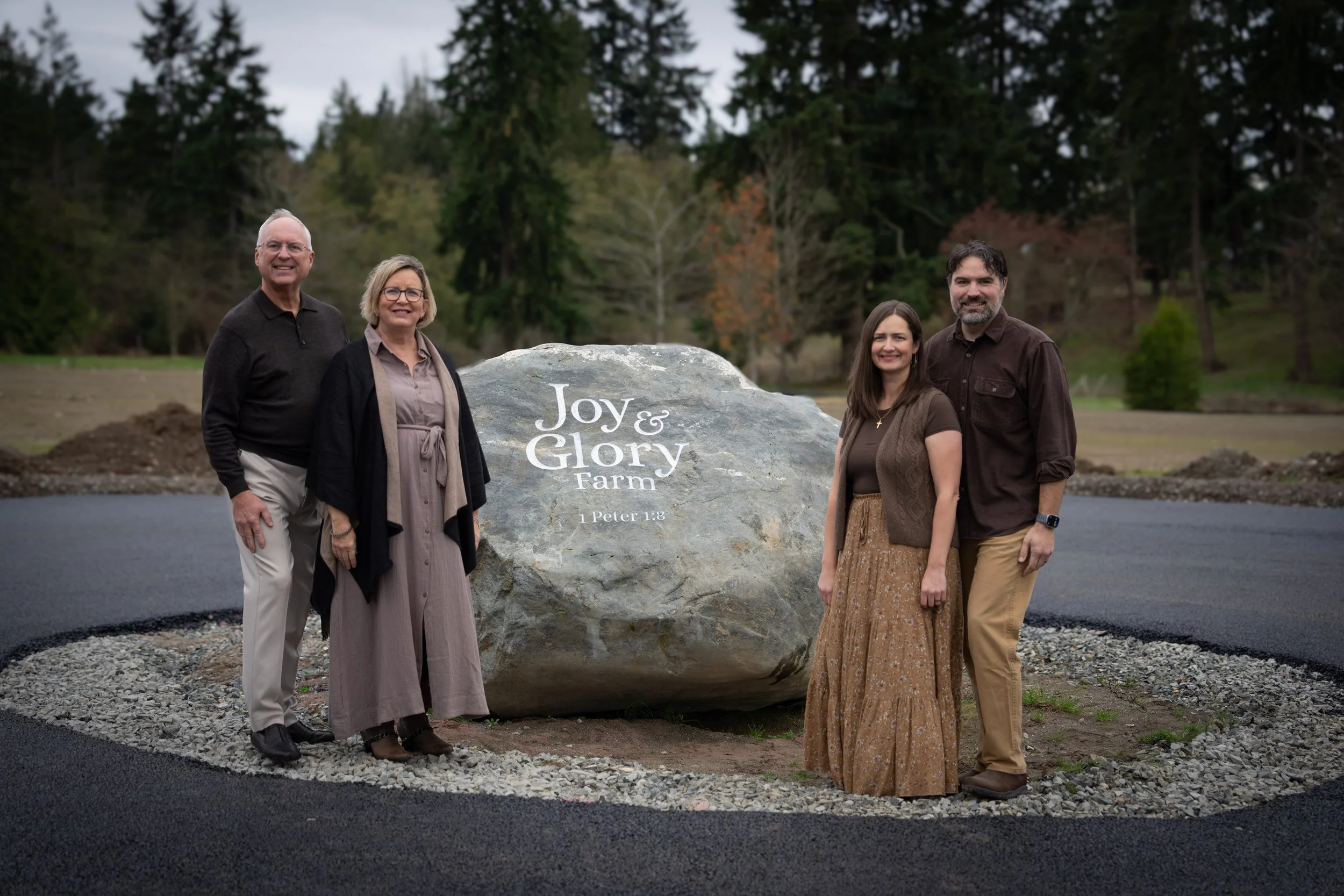 A family of five standing outdoors on a paved area behind a large stone. The stone has the words "Joy & Glory Farm" and "1 Peter 1:8" inscribed on it. The background features trees and a cloudy sky.